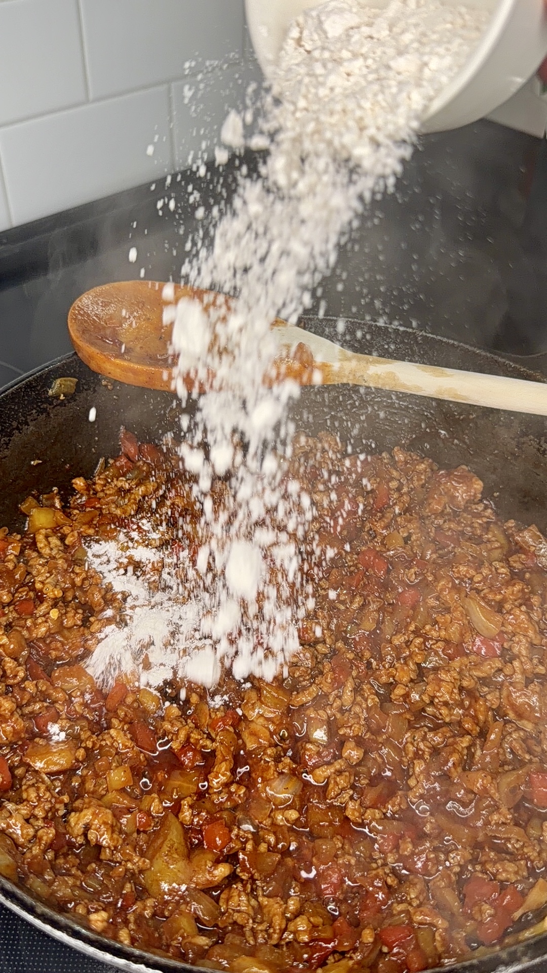 A hand pours flour into a skillet filled with sizzling ground meat, chopped vegetables, and sauce on a stovetop. A wooden spoon rests across the pan.