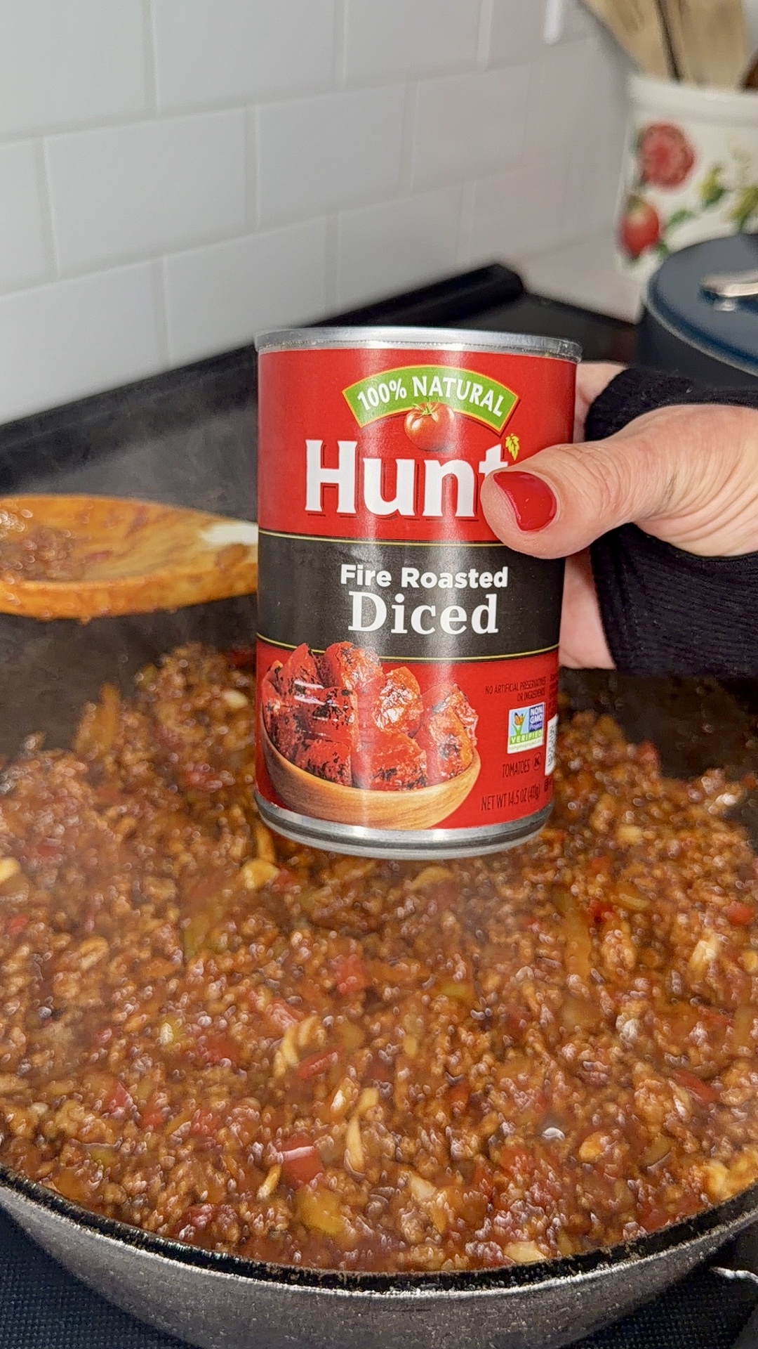 A hand holds a can of Hunt’s Fire Roasted Diced tomatoes over a pan of simmering meat sauce, with a wooden spoon stirring the mixture.