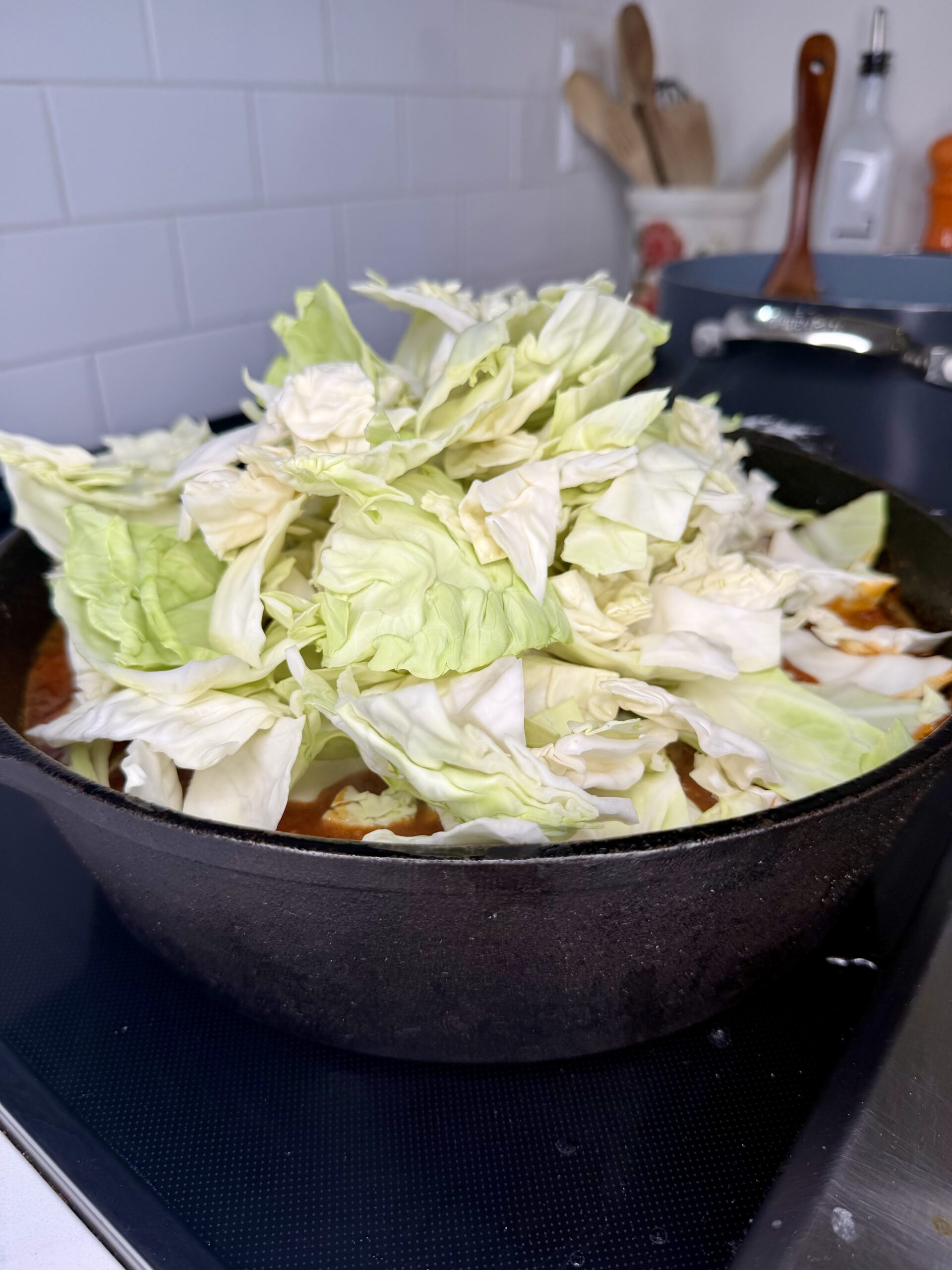 A cast iron skillet filled with chopped cabbage sits on a stovetop, surrounded by kitchen utensils and white tile backsplash in the background.