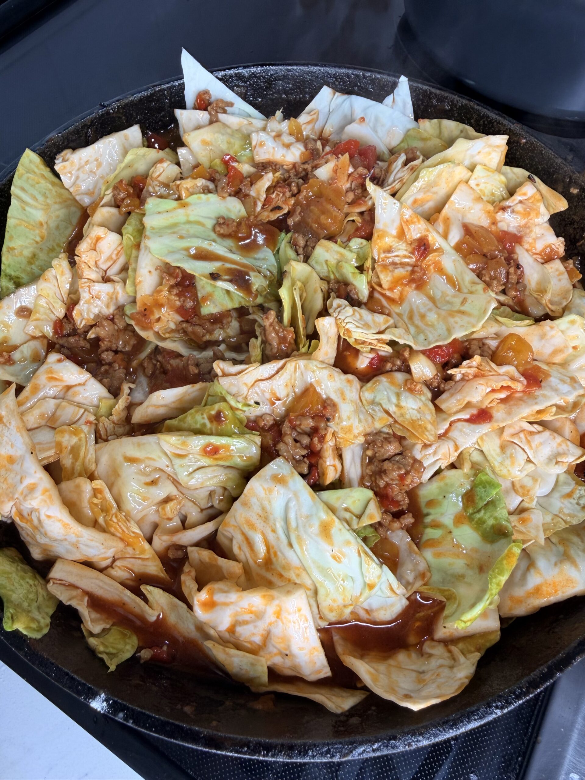 A skillet filled with chopped cabbage, ground meat, tomatoes, and sauce, all cooking together. The dish appears colorful with green, white, and red tones, and is being prepared on a stovetop.