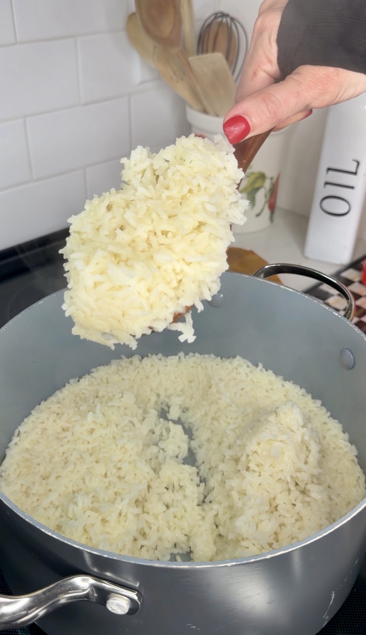 A hand holds a spoonful of cooked white rice above a pot filled with more rice. A white container labeled OIL is visible in the background.