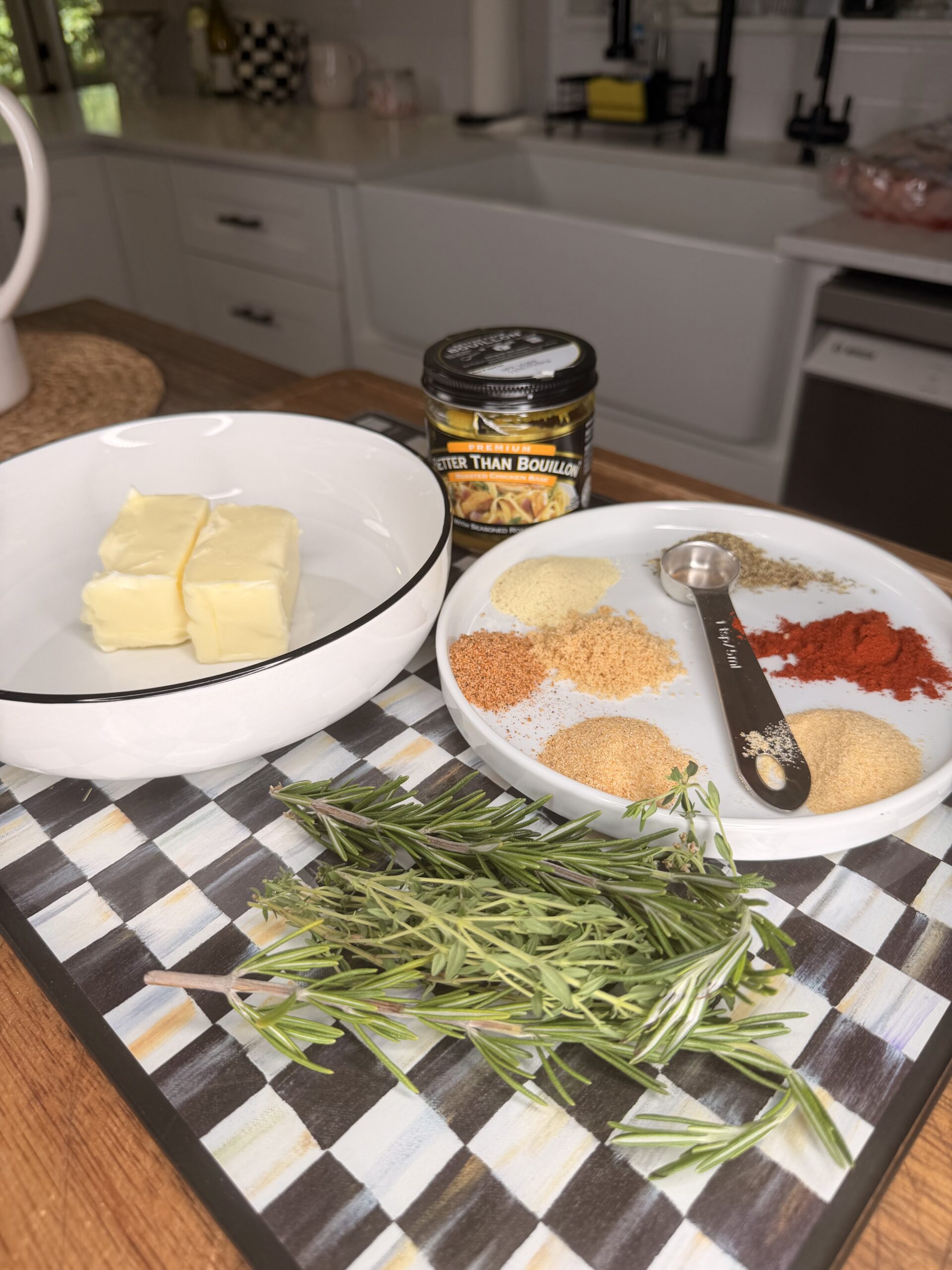 Butter in a bowl, a jar of bouillon, a plate of various spices with a measuring spoon, and fresh rosemary sprigs on a checkered cutting board in a bright kitchen.