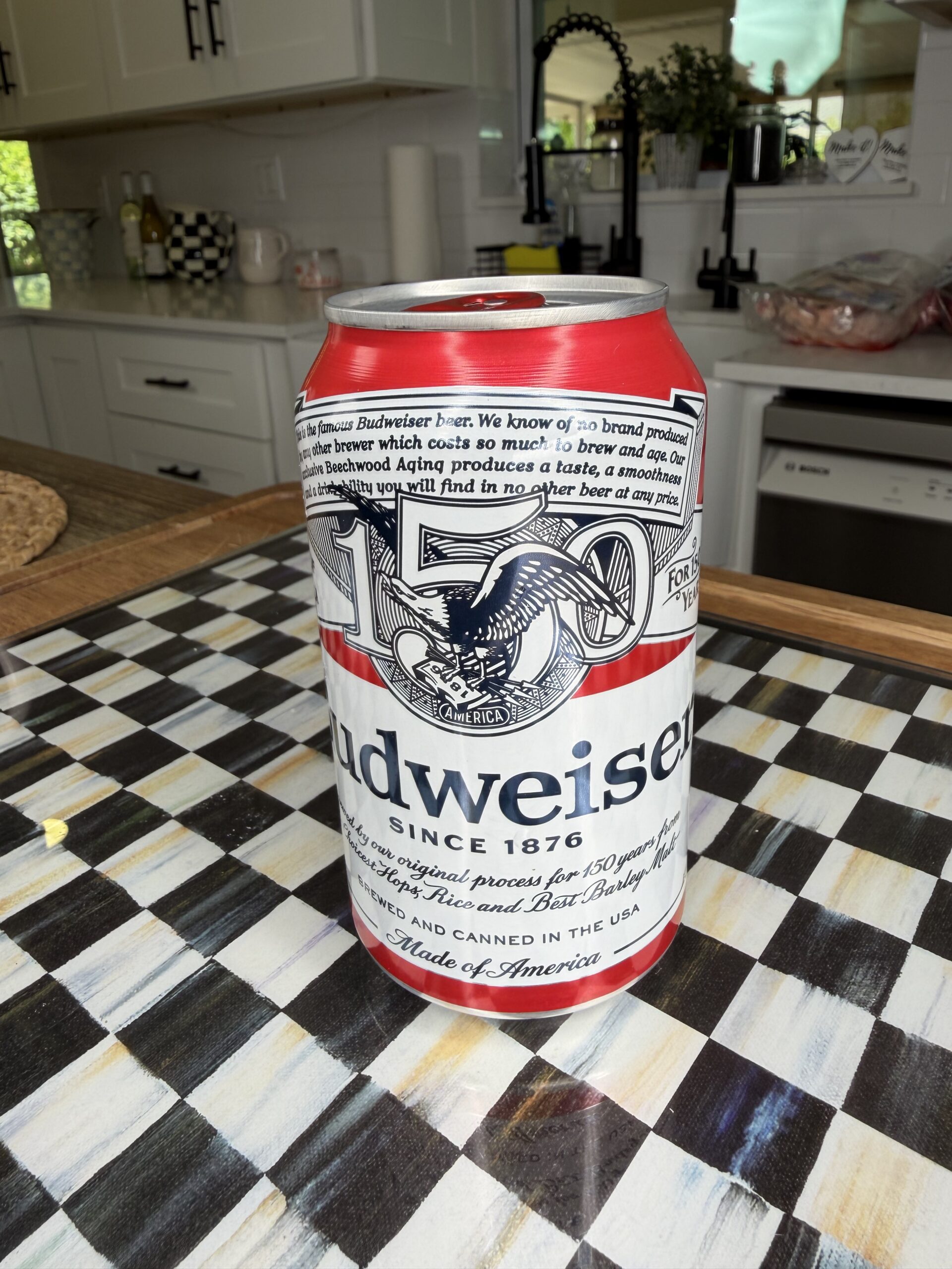 A Budweiser beer can is placed on a checkered countertop in a bright kitchen with white cabinets and various kitchen items in the background.