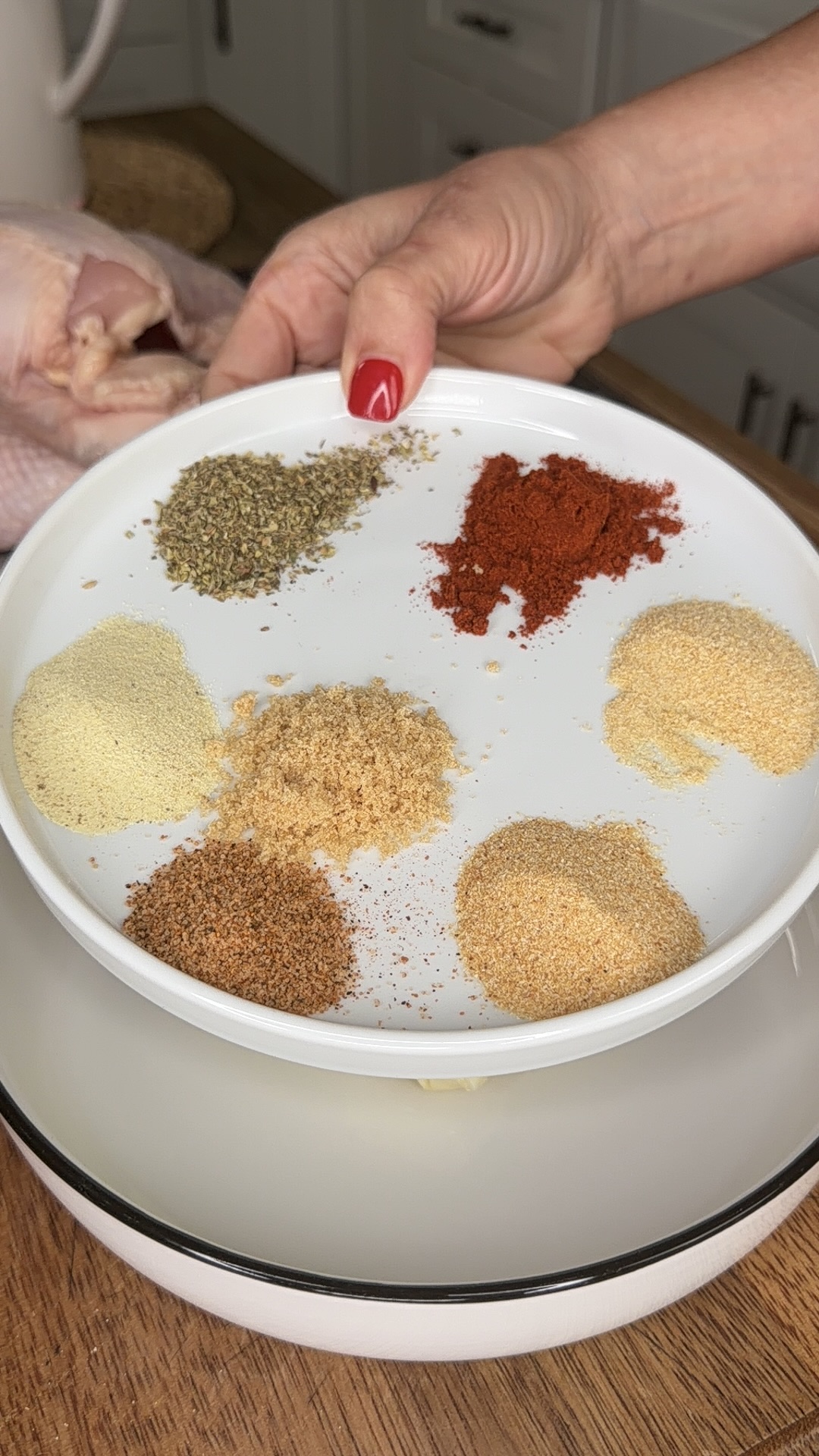 A hand with red nail polish holds a white plate with six piles of spices, next to a raw chicken on a wooden surface in a kitchen.