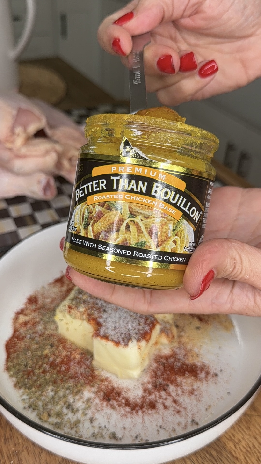 A hand with red nails scoops Better Than Bouillon roasted chicken base from a jar over a bowl with butter, spices, and herbs. A raw chicken and checkered cloth are visible in the background.