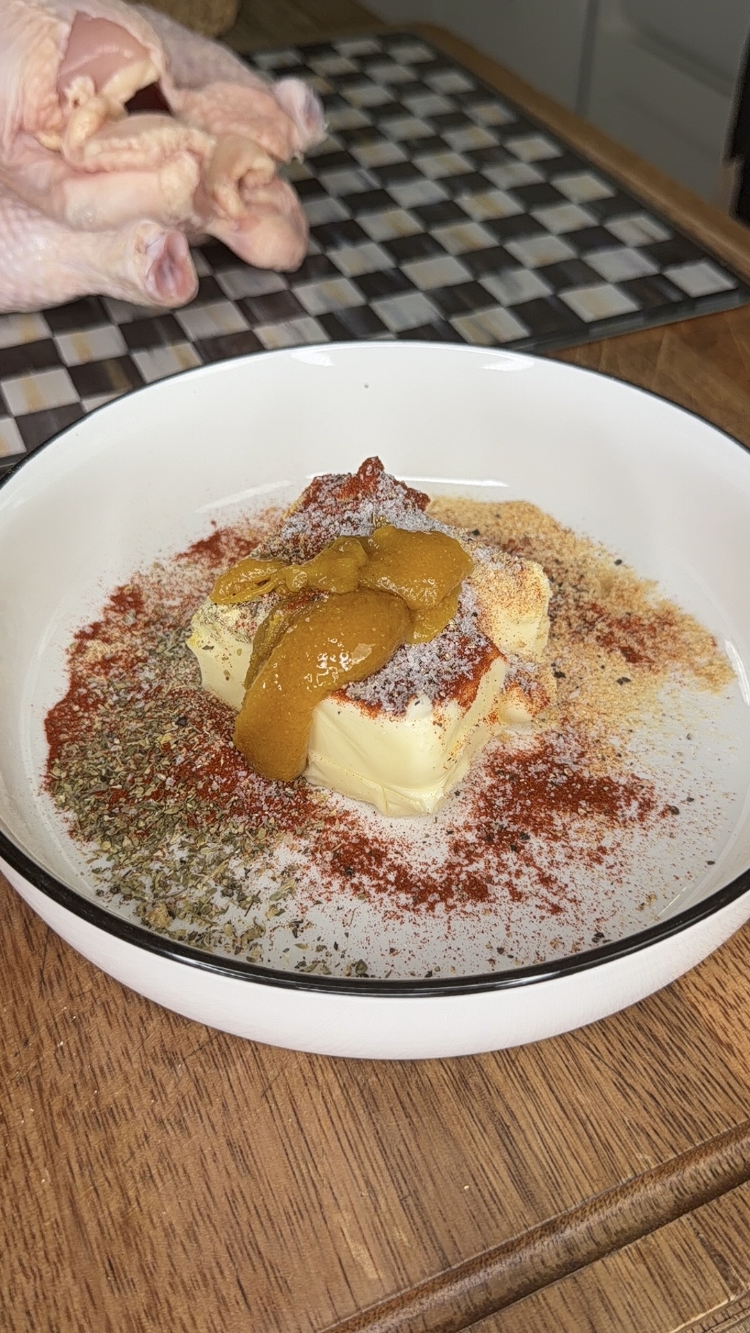 A bowl with a stick of butter topped with spices and mustard sits on a wooden counter; raw chicken is visible nearby on a cutting board.