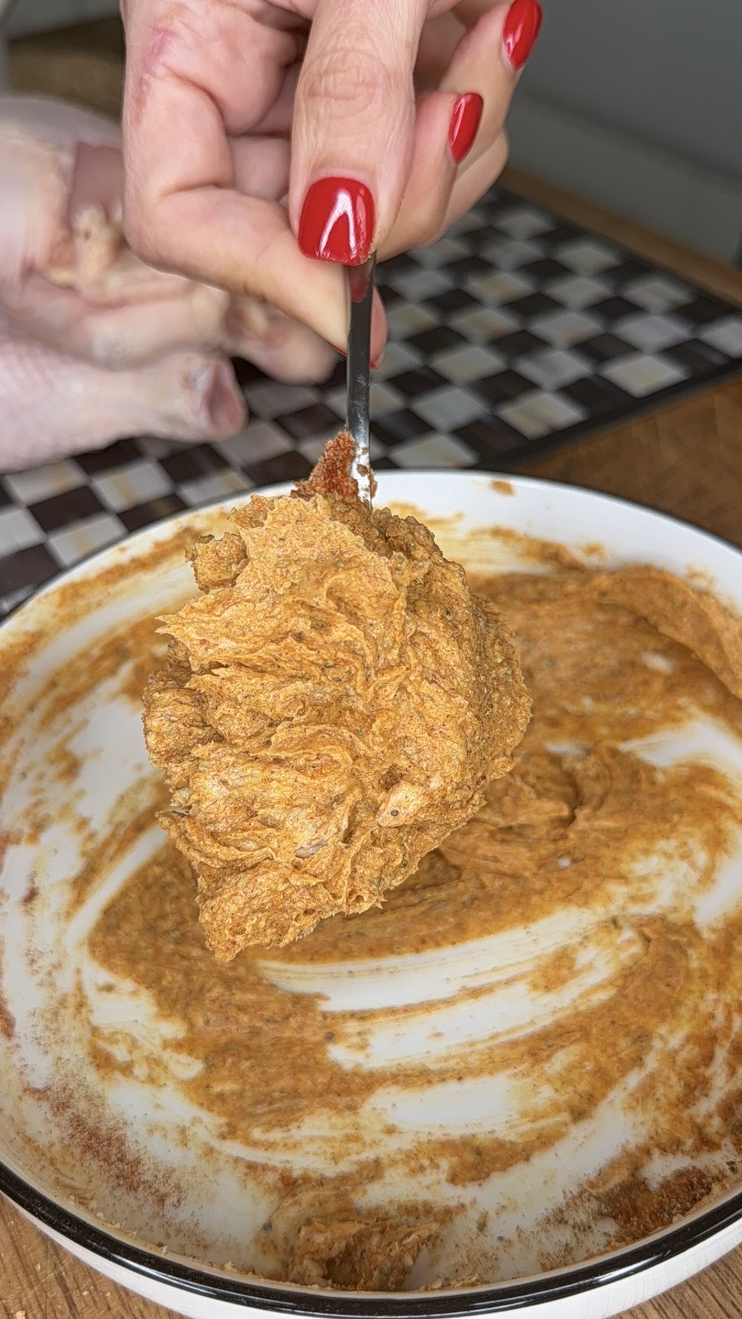 A hand with red nail polish holds a spoonful of seasoned paste over a bowl, with a raw chicken and a checkered cutting board in the background.