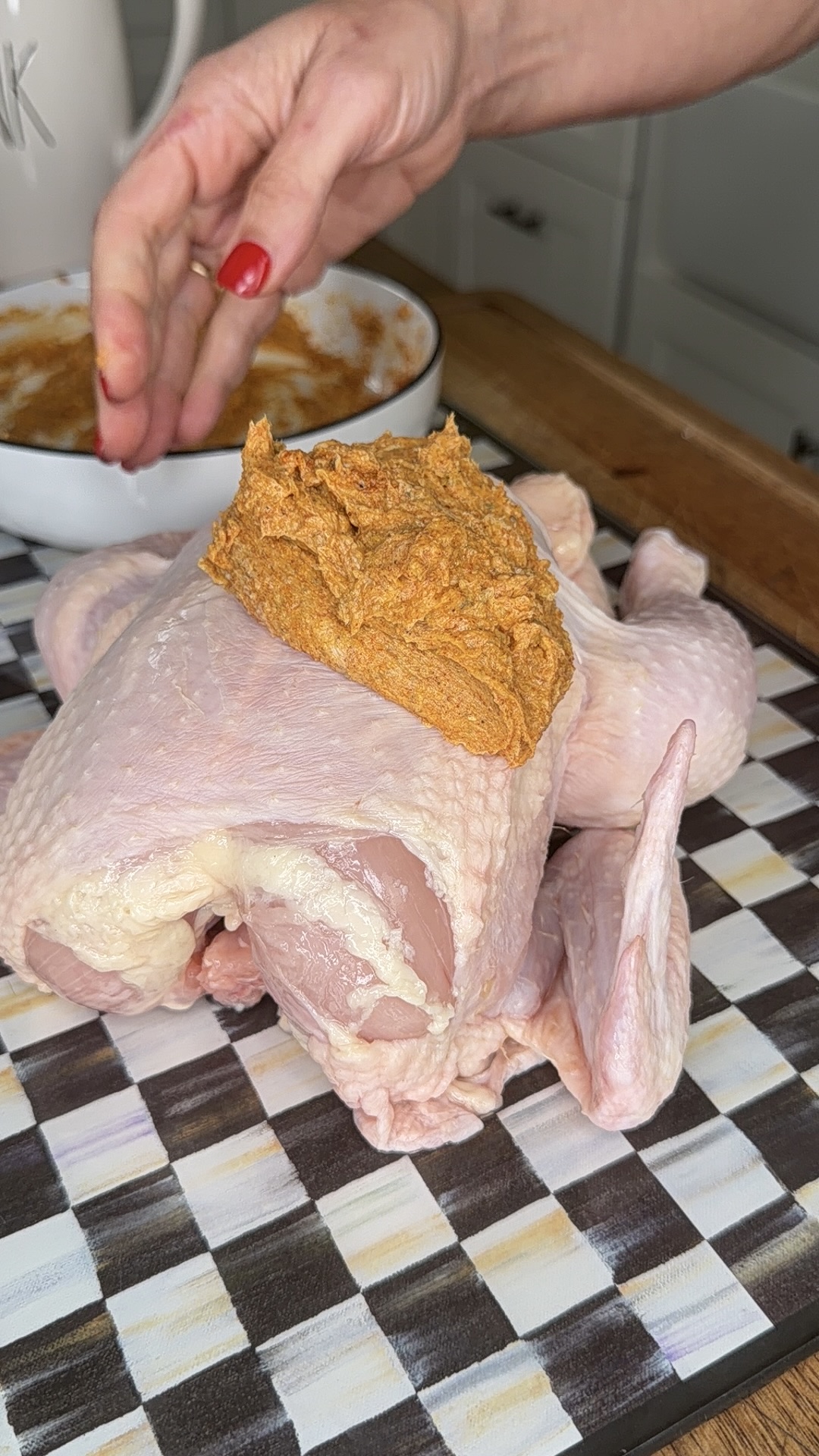 A hand with red nail polish spreads a thick orange spice paste onto a raw whole chicken on a checkered cutting board in a kitchen.