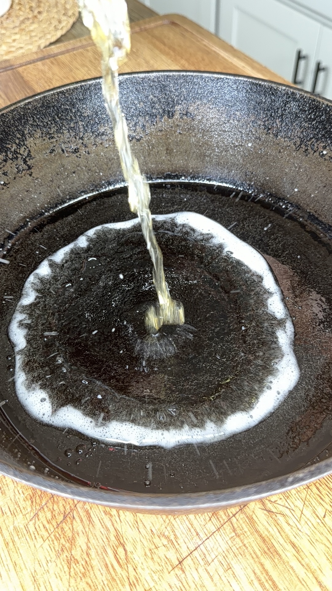 Liquid is being poured into a hot, black cast iron skillet on a wooden countertop, creating bubbles and foam in the center of the pan.