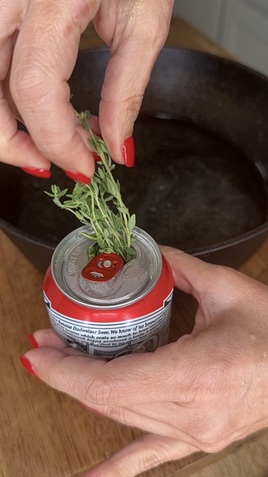 A person with red nail polish places fresh herbs into the opening of a red soda can, with a cast iron pan filled with liquid in the background.