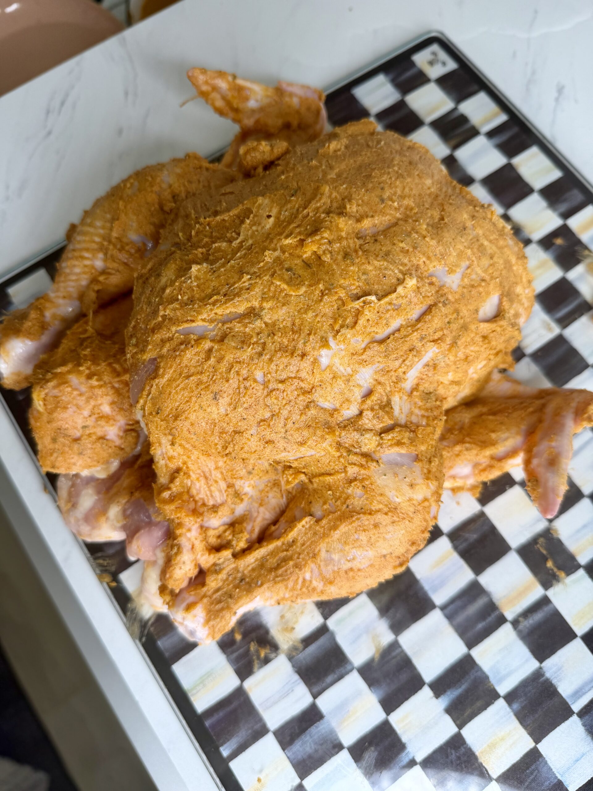 A whole raw chicken coated in orange-colored marinade sits on a black and white checkered tray on a light-colored countertop.