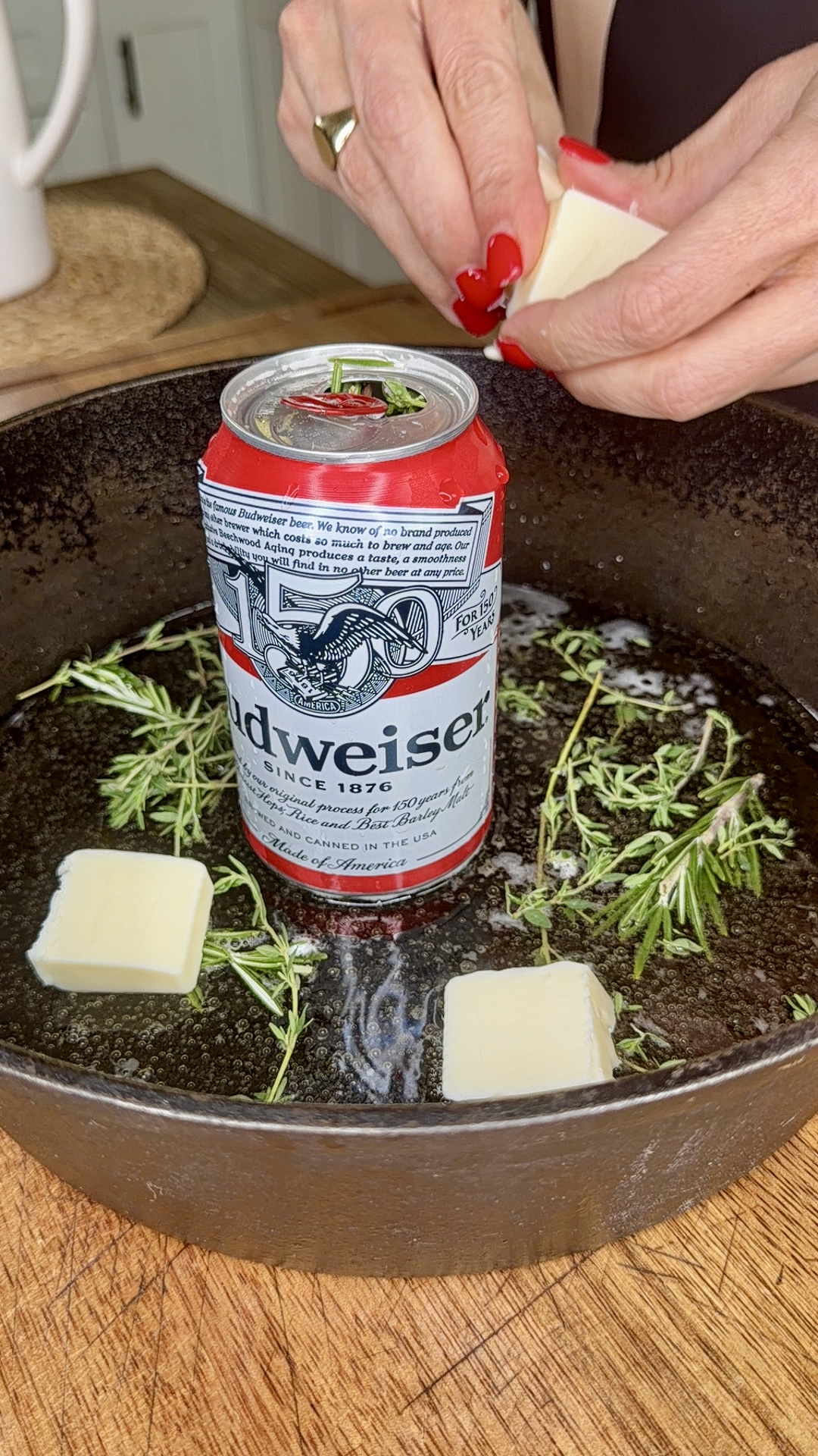 A can of Budweiser stands in a cast iron skillet with melted butter, rosemary, and butter cubes. A person with red nails adds another butter cube.
