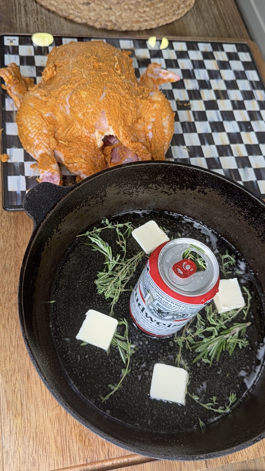 A raw seasoned chicken on a cutting board sits next to a cast iron skillet containing a beer can, butter cubes, rosemary, and garlic cloves, ready for cooking.