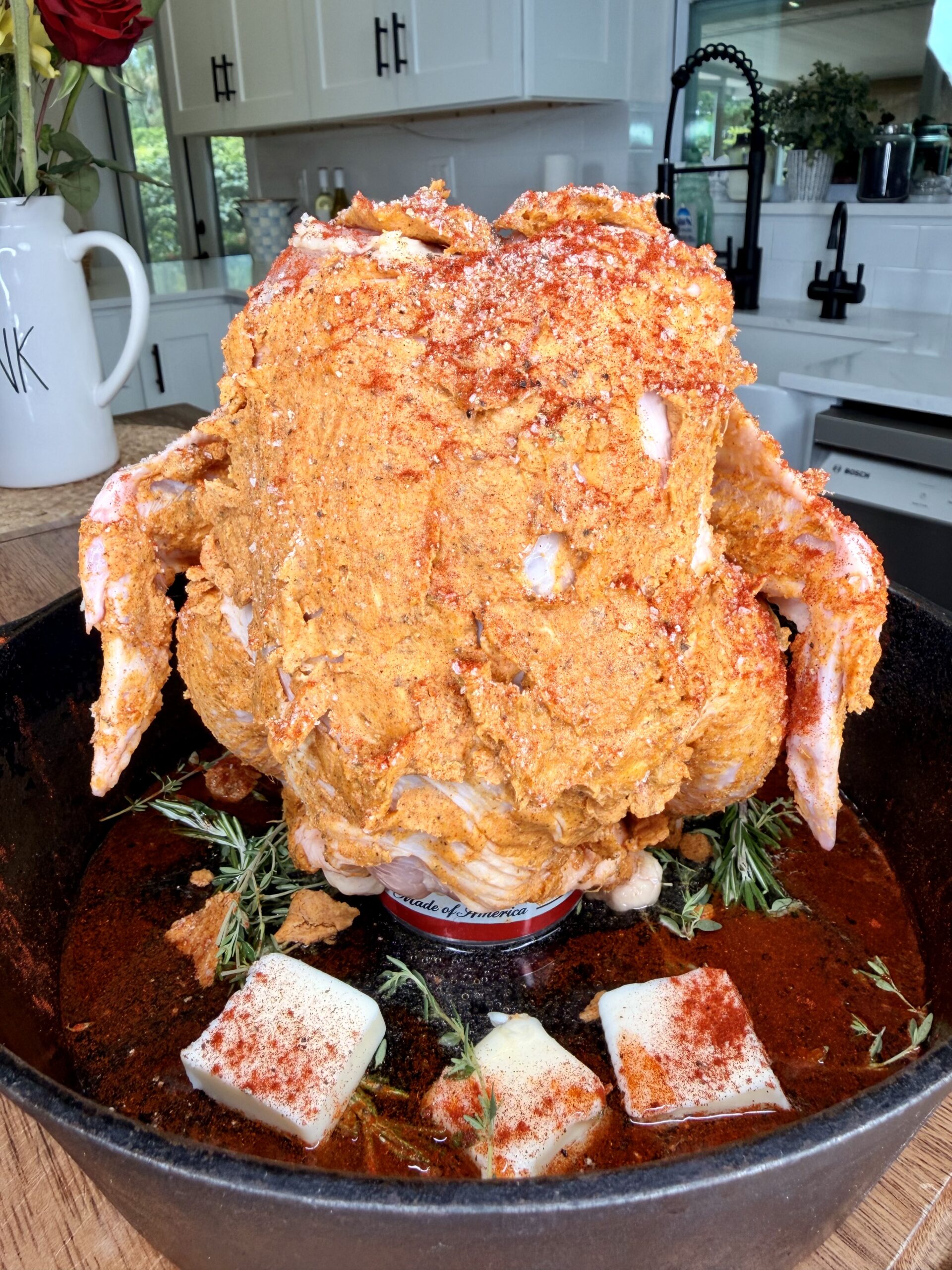 A whole raw chicken coated in seasoning sits upright on a can in a cast iron pan with herbs, butter cubes, and spices, ready to be cooked in a bright kitchen.