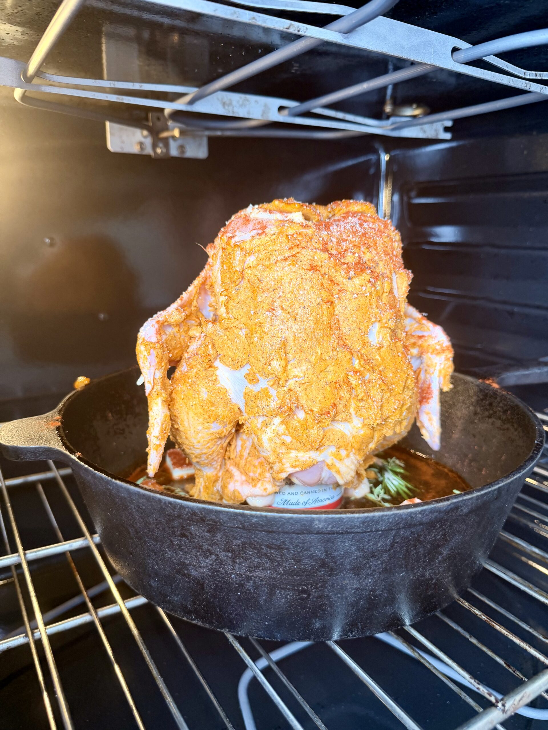 A whole seasoned chicken sits upright on a beer can in a cast iron skillet, placed inside an oven, ready to be roasted.