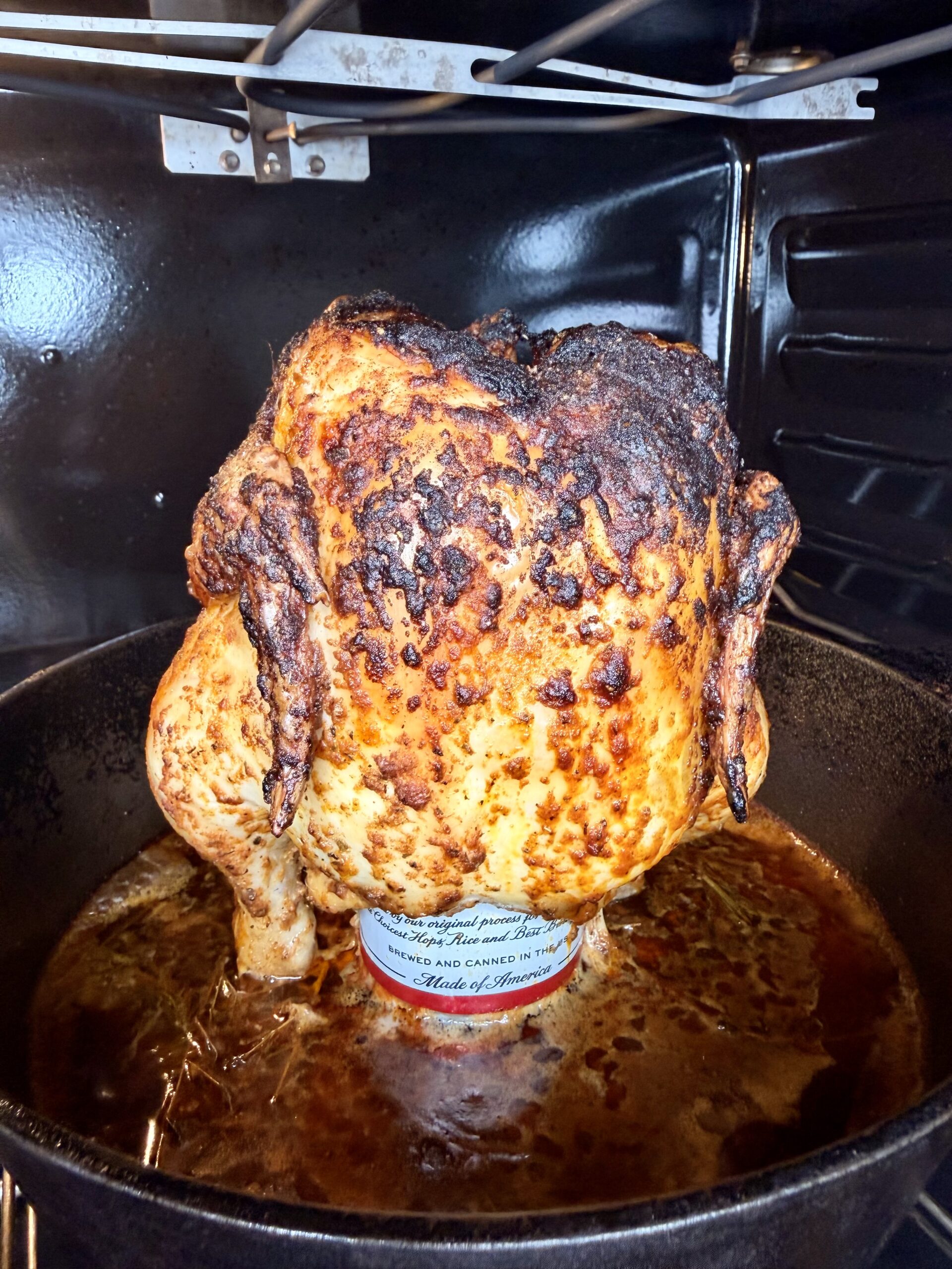 A whole roasted chicken sits upright on a can inside a cast iron pan in an oven, with juices collecting in the pan and crispy, browned skin on the chicken.