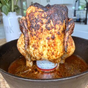 A whole roasted chicken is upright in a cast iron skillet, sitting on a can, with a charred, seasoned skin. Kitchen background with flowers visible.