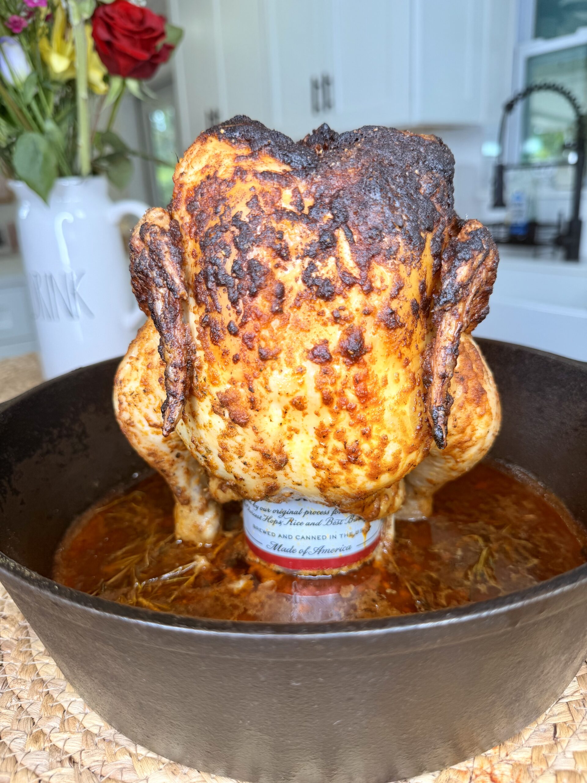 A whole roasted chicken is upright in a cast iron skillet, sitting on a can, with a charred, seasoned skin. Kitchen background with flowers visible.