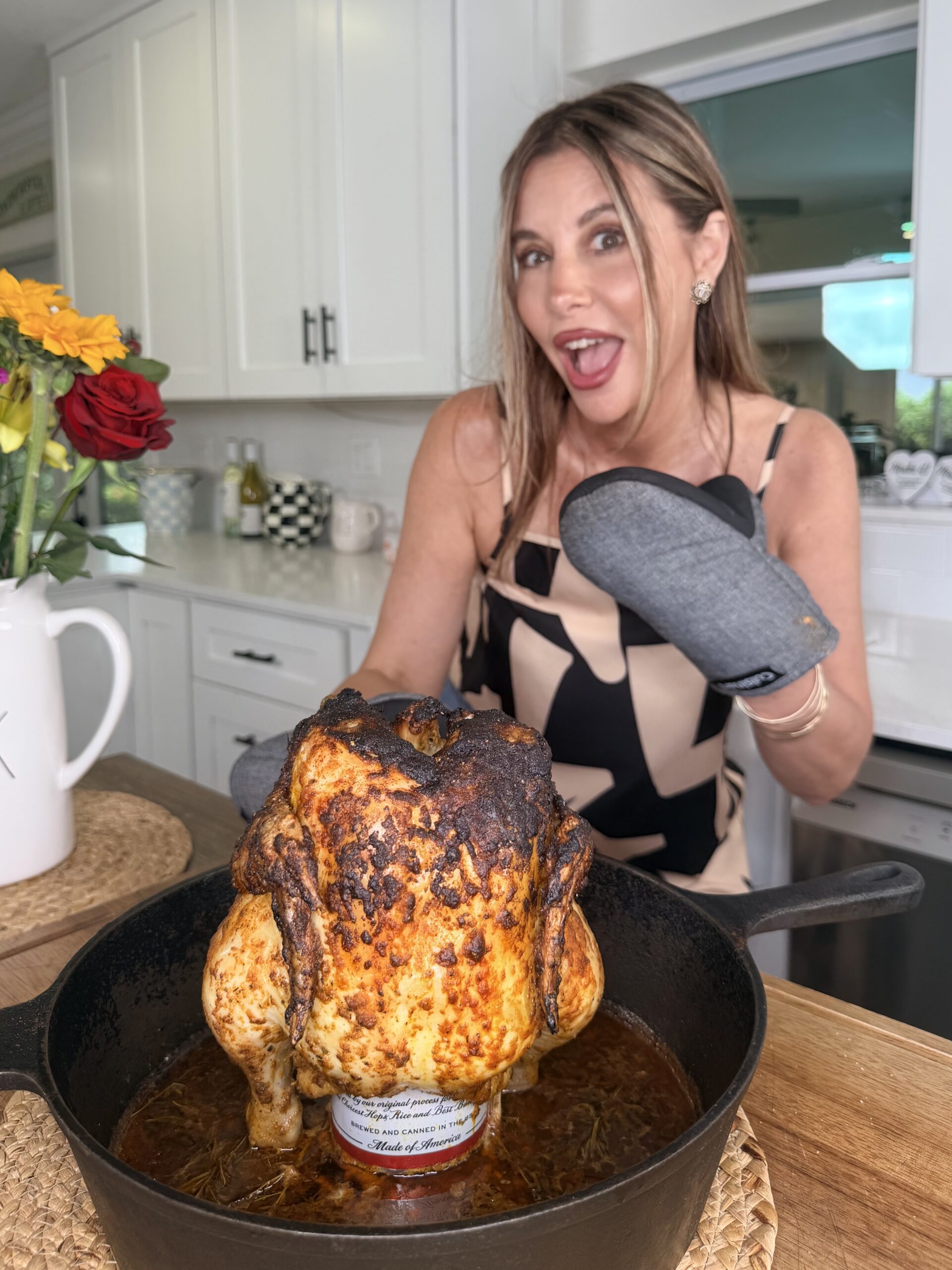 A woman in a kitchen, wearing an oven mitt, smiles excitedly behind a roasted chicken cooked upright in a cast iron skillet on top of a can.