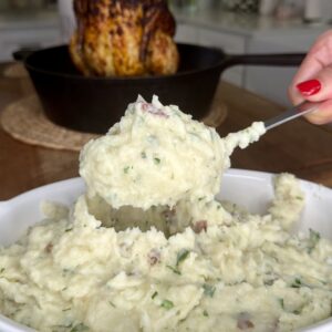 A hand with red nail polish scoops creamy mashed potatoes with herbs from a white dish. In the background, a roasted chicken sits in a cast iron skillet.
