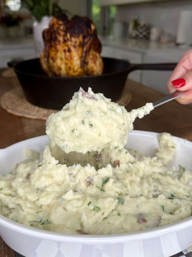 A hand with red nail polish scoops creamy mashed potatoes with herbs from a white dish. In the background, a roasted chicken sits in a cast iron skillet.