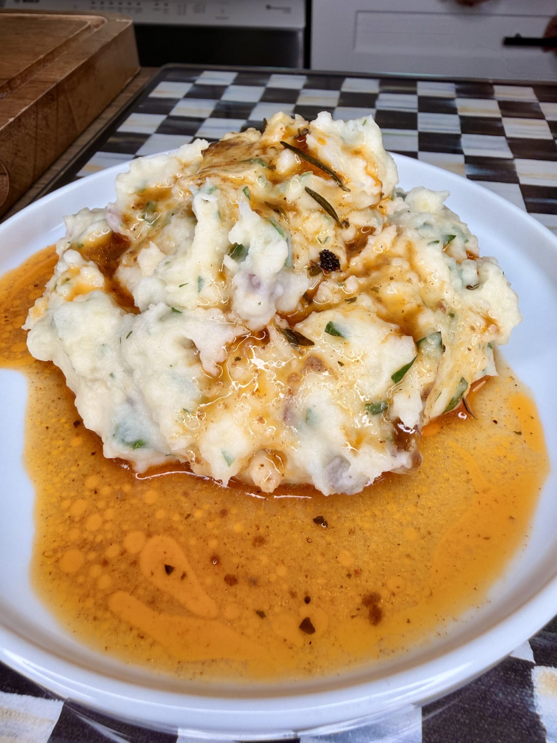 A plate of creamy mashed potatoes mixed with herbs, topped with a generous drizzle of reddish-brown gravy or spiced oil, sits on a white plate over a black-and-white checkered surface.