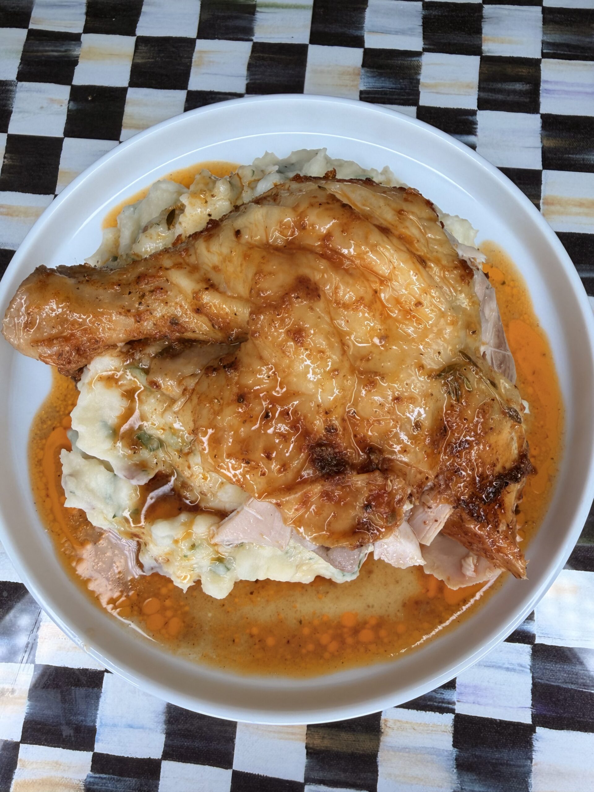 A roasted chicken leg sits on mashed potatoes in a white bowl, with orange-colored gravy pooling around it. The dish is placed on a checkered black, white, and gold table.