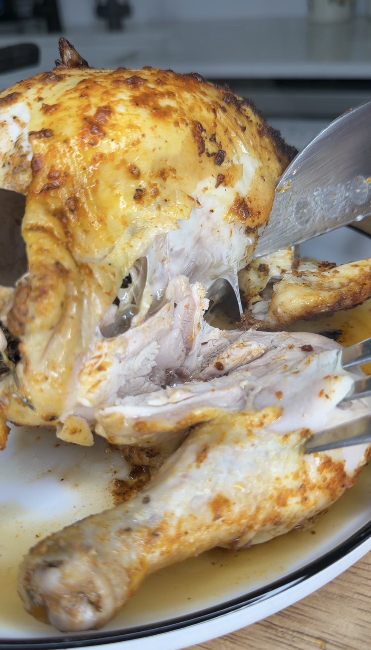A close-up of a roasted chicken being carved with a knife and fork, showing juicy, tender white and dark meat inside, on a white plate.