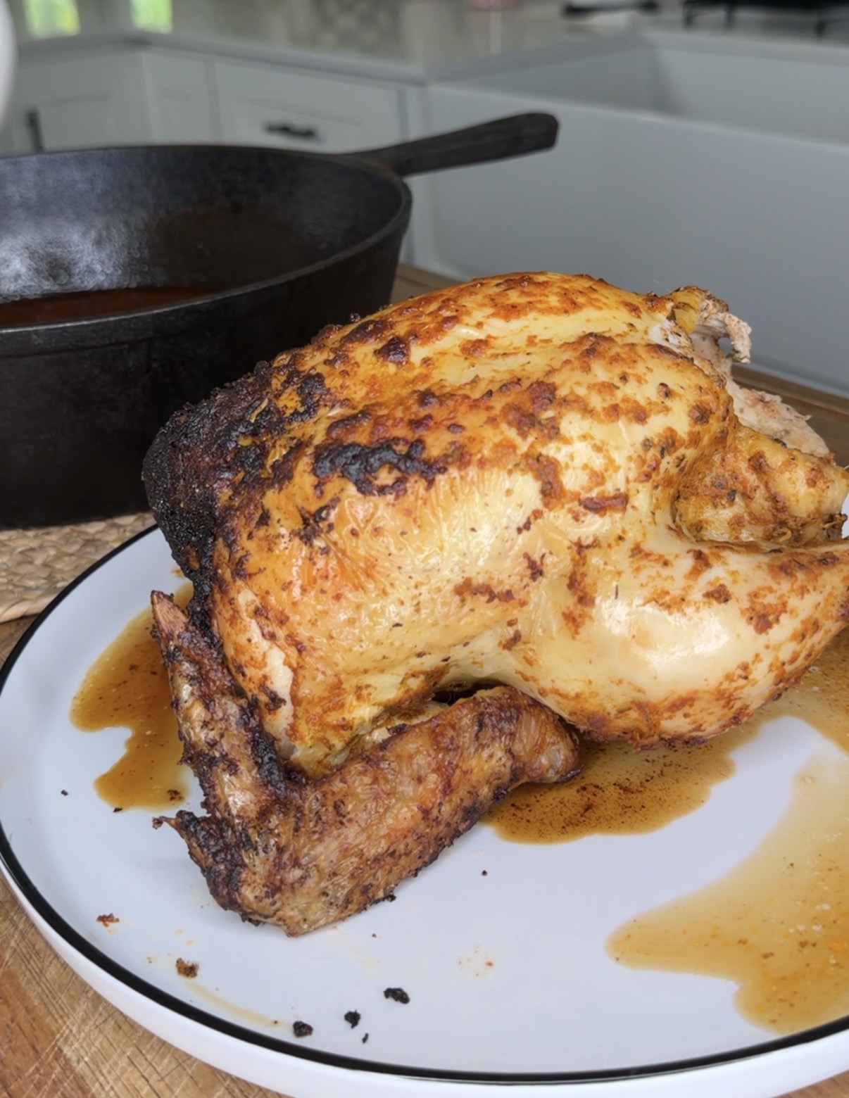 A whole roasted chicken with crispy, golden skin sits on a white plate, with juices pooling beneath it. A black cast iron skillet is visible in the background on a kitchen counter.