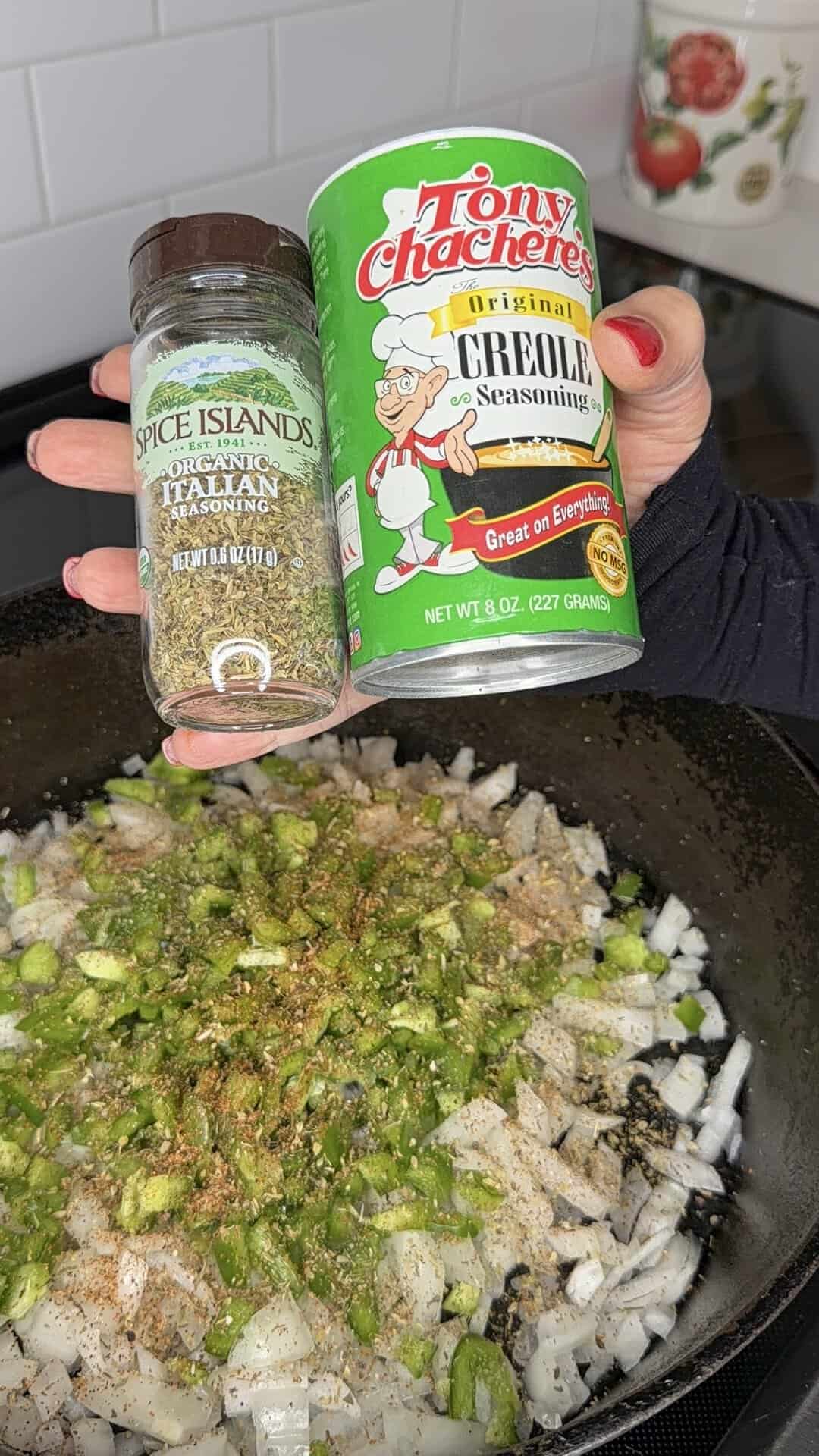 A hand holds Italian seasoning and Creole seasoning above diced onions and green peppers cooking in a skillet.