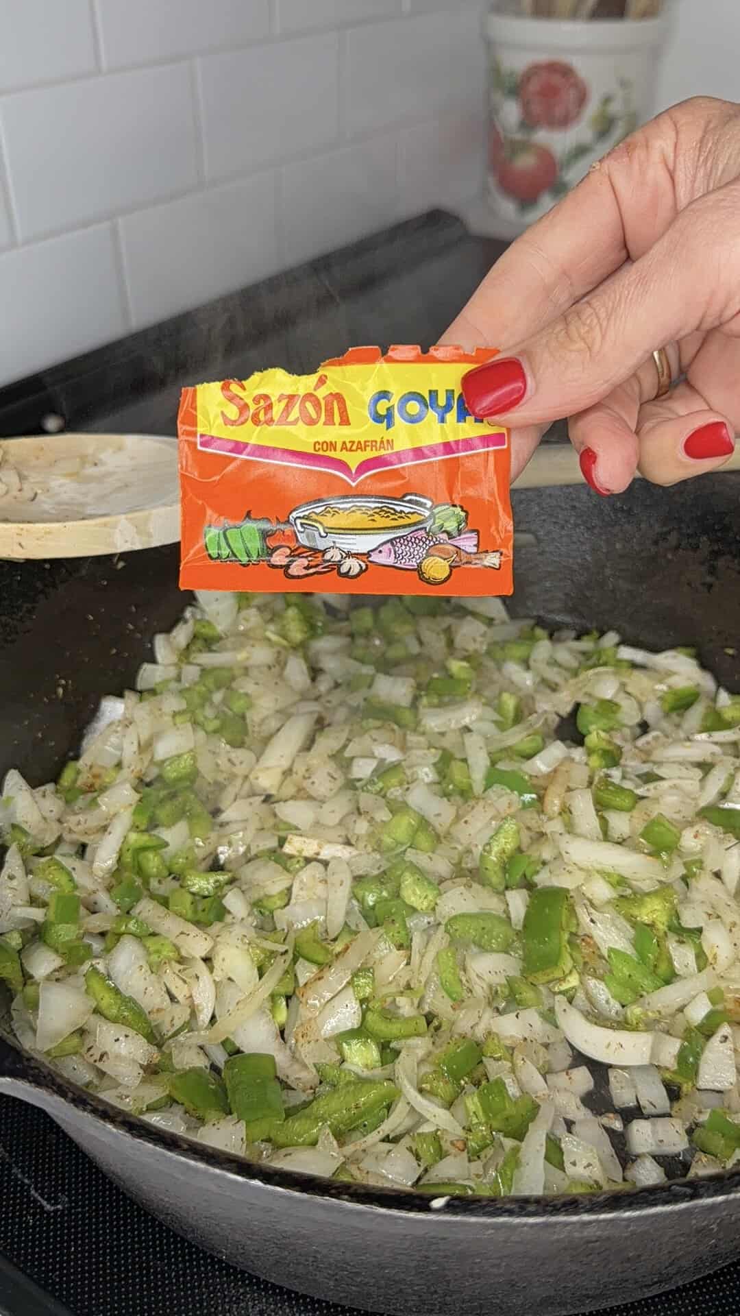 A hand holds a Sazon Goya seasoning packet over a skillet with chopped onions and green peppers being sautéed. A wooden spoon stirs the vegetables in the black pan.