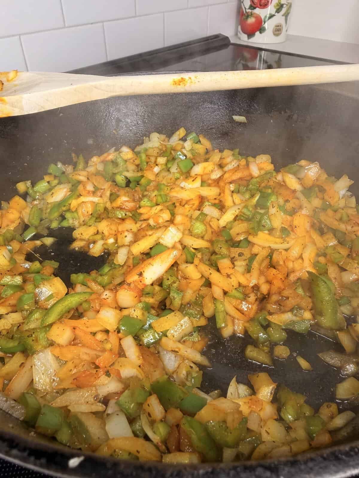 Diced onions, green bell peppers, and orange bell peppers are being sautéed with spices in a black skillet. A wooden spoon rests on the pan’s edge, and steam is rising from the vegetables.