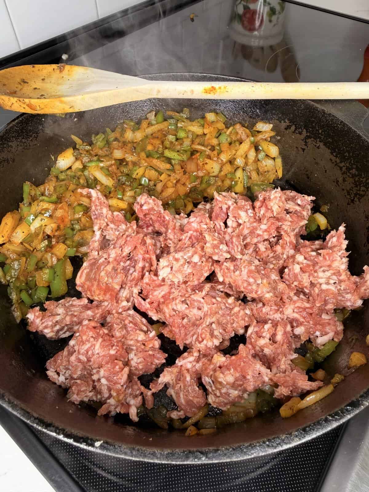 Ground meat and sautéed onions, bell peppers, and spices are cooking together in a black cast iron pan on a stovetop. A wooden spoon rests on the edge of the pan. Steam is visible.