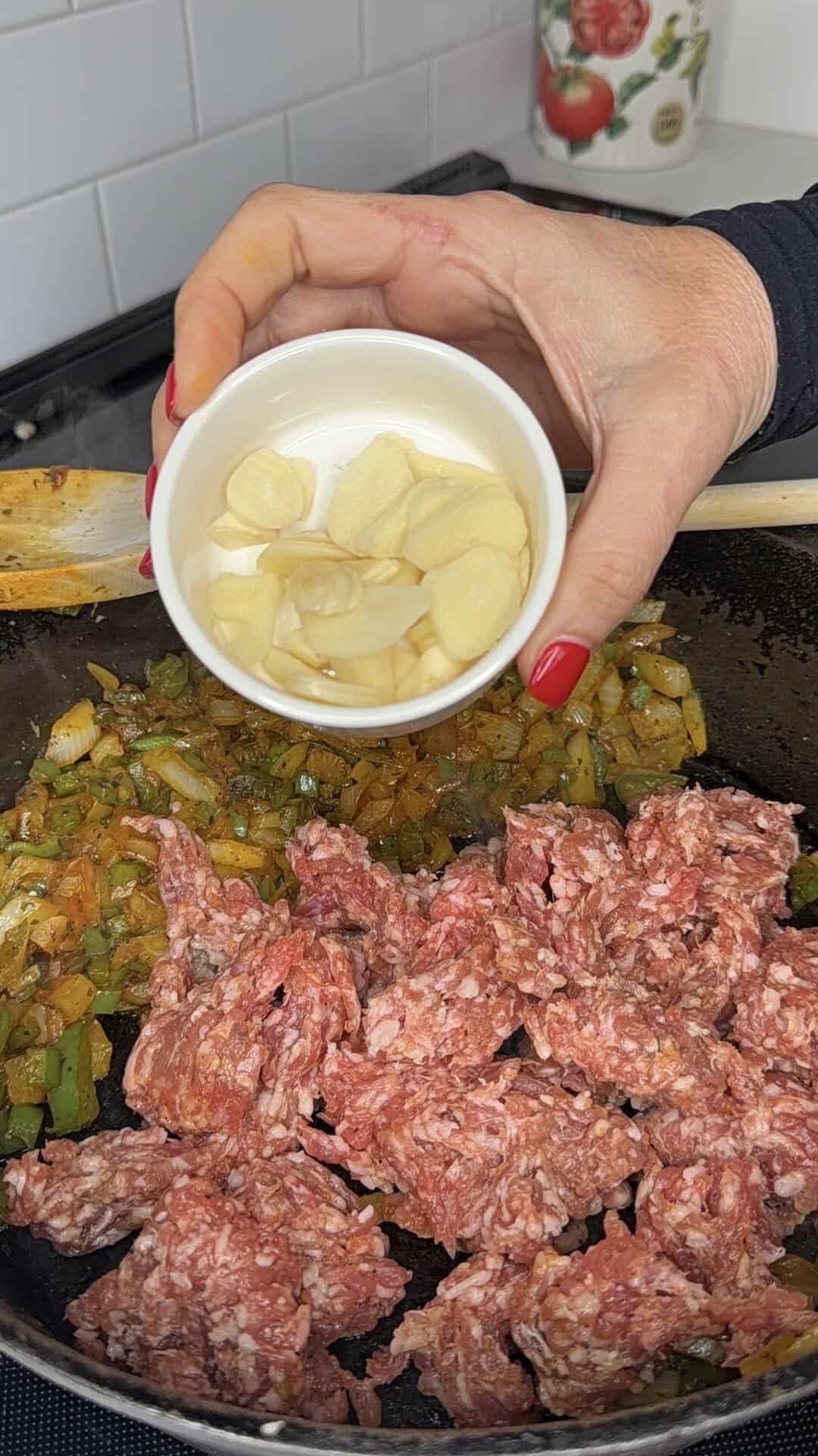 A hand holds a small bowl of sliced garlic over a skillet containing chopped onions, bell peppers, and raw ground meat on a stovetop.