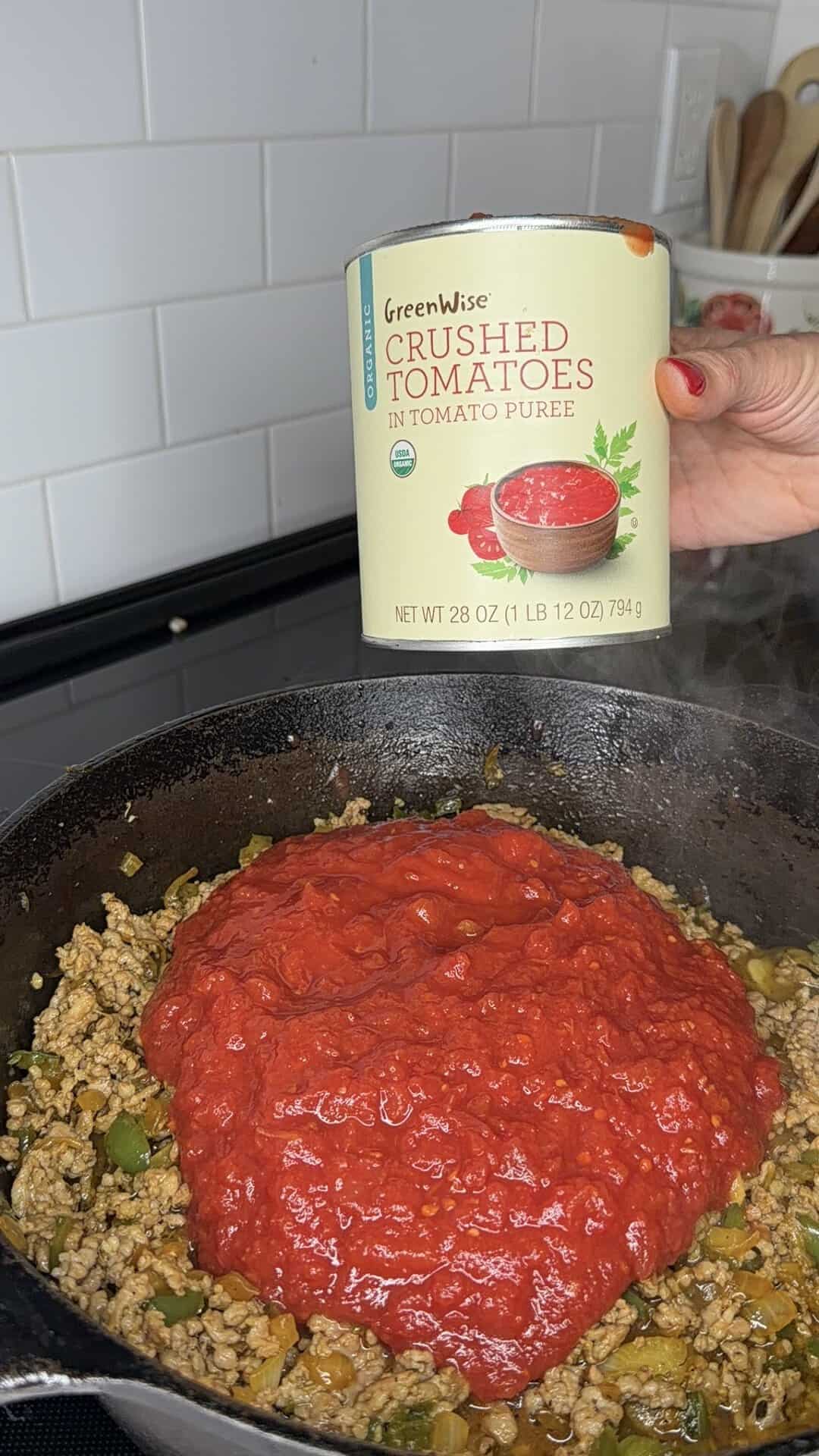 A hand holds a can of GreenWise crushed tomatoes above a skillet filled with ground meat, vegetables, and tomato sauce on a stovetop.