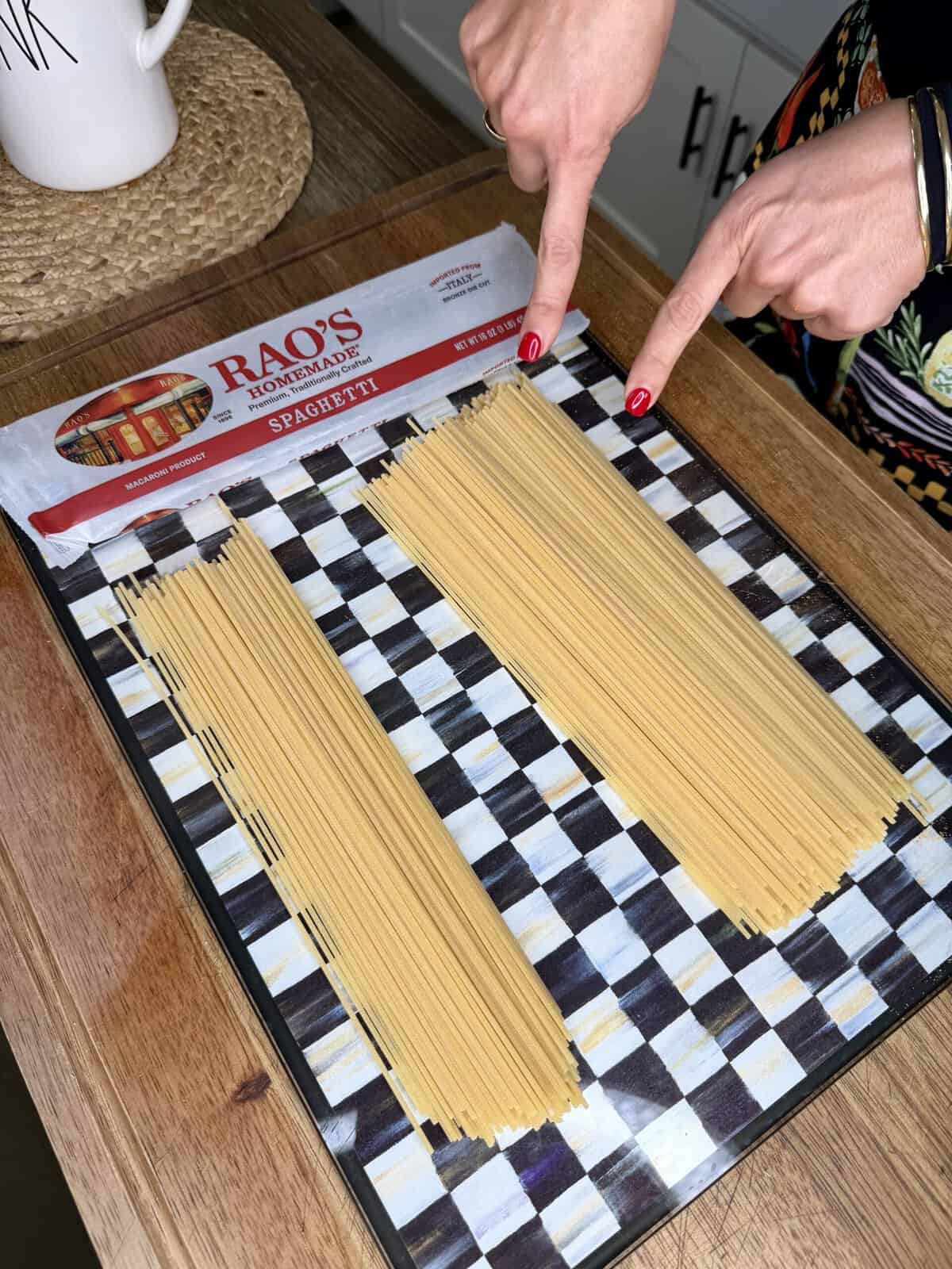 Two hands point at two piles of uncooked spaghetti on a checkered cutting board next to a Rao’s Homemade Spaghetti package on a wooden counter.