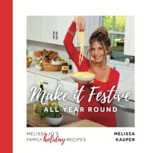 A smiling woman in a red dress stands in a kitchen, holding a plate of pasta and lifting noodles with one hand. The text reads Make it Festive All Year Round and Melissa Jos Family Holiday Recipes by Melissa Kauper.