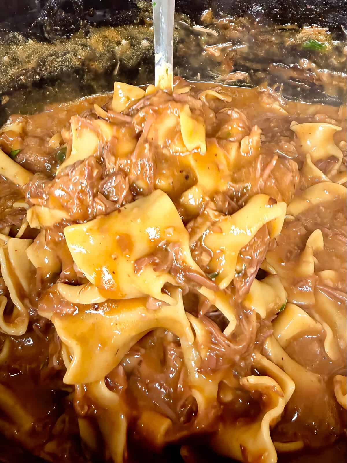 Wide, flat noodles topped with a rich, brown, saucy beef mixture, being stirred in a pan. The dish appears hearty and savory, with visible chunks of beef and sauce coating the noodles.
