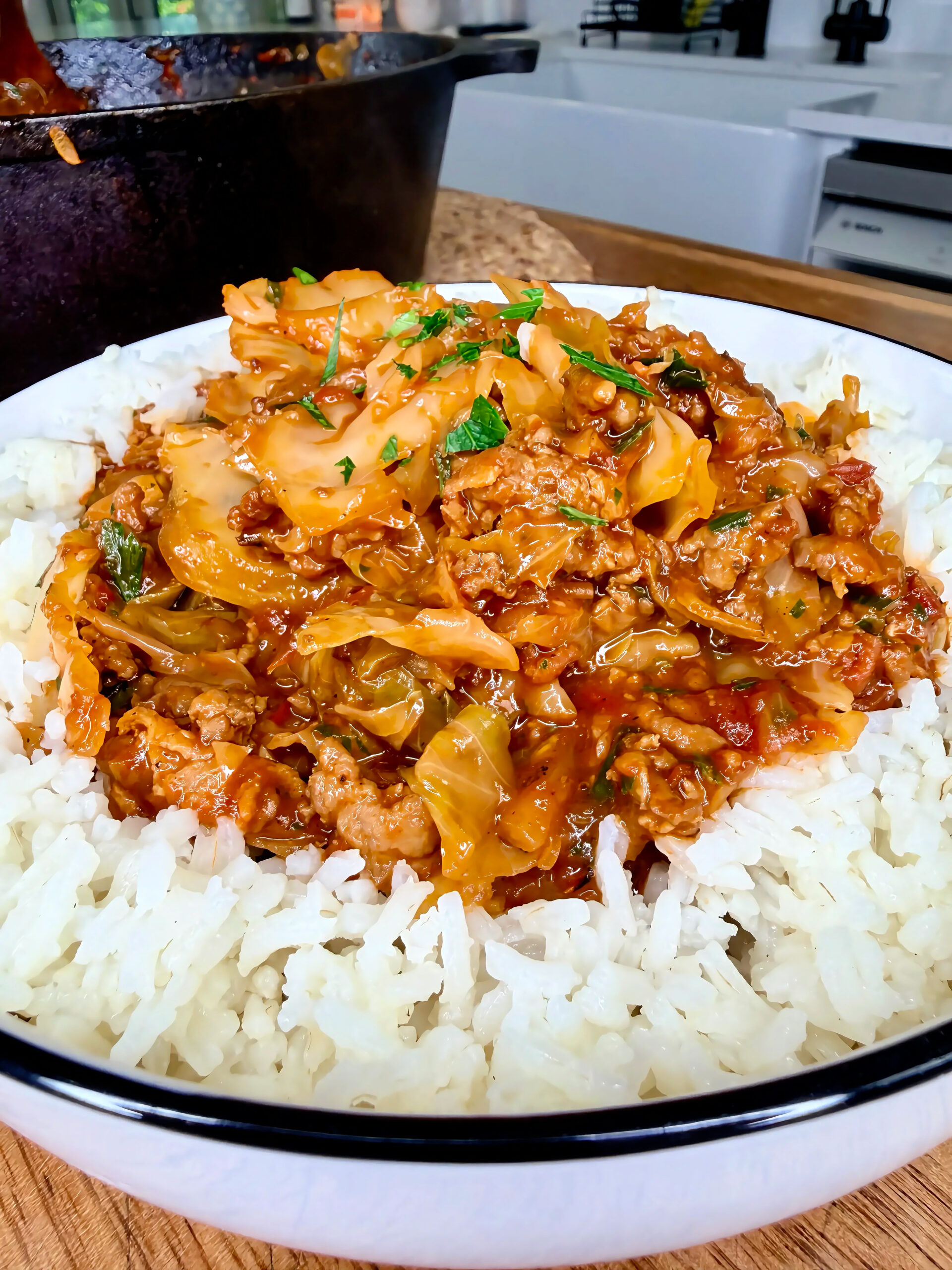 A white bowl filled with steamed white rice topped with saucy ground meat, cabbage, and herbs. The background shows a kitchen and a cooking pot.