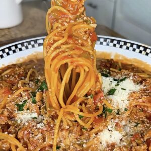 A fork lifts a swirl of spaghetti with meat sauce from a plate, topped with grated cheese and chopped herbs, in a kitchen setting.