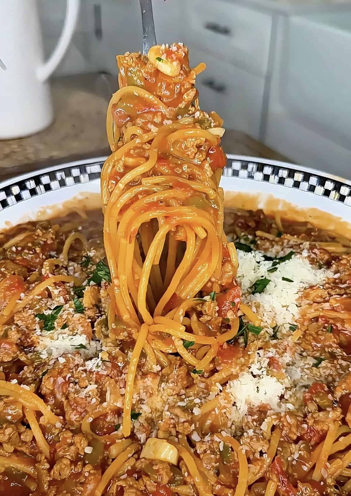 A fork lifts a swirl of spaghetti with meat sauce from a plate, topped with grated cheese and chopped herbs, in a kitchen setting.