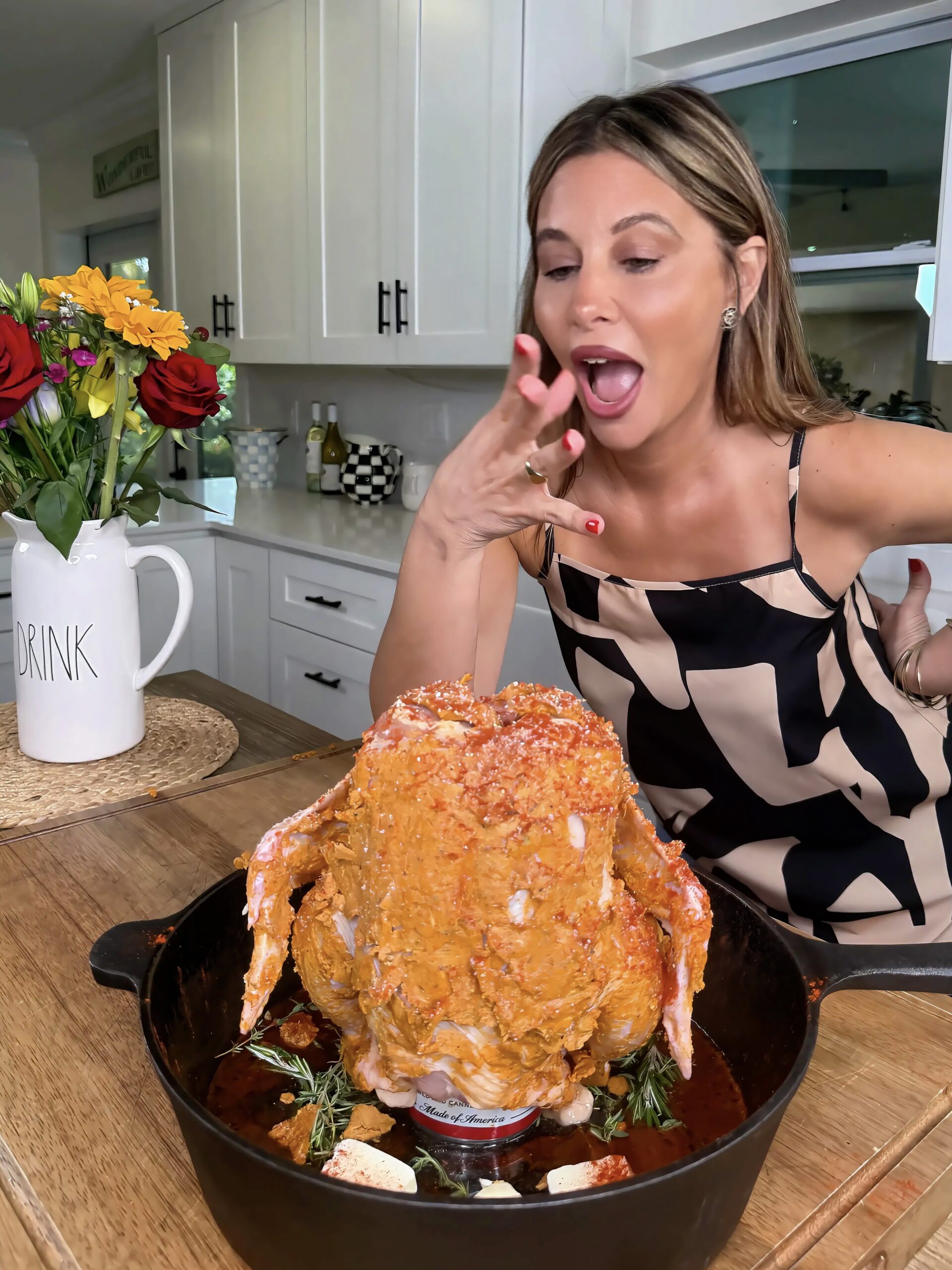 A woman in a patterned dress smiles and gestures excitedly toward a seasoned whole chicken upright in a pot, ready to be cooked in a kitchen with white cabinets and a vase of flowers.