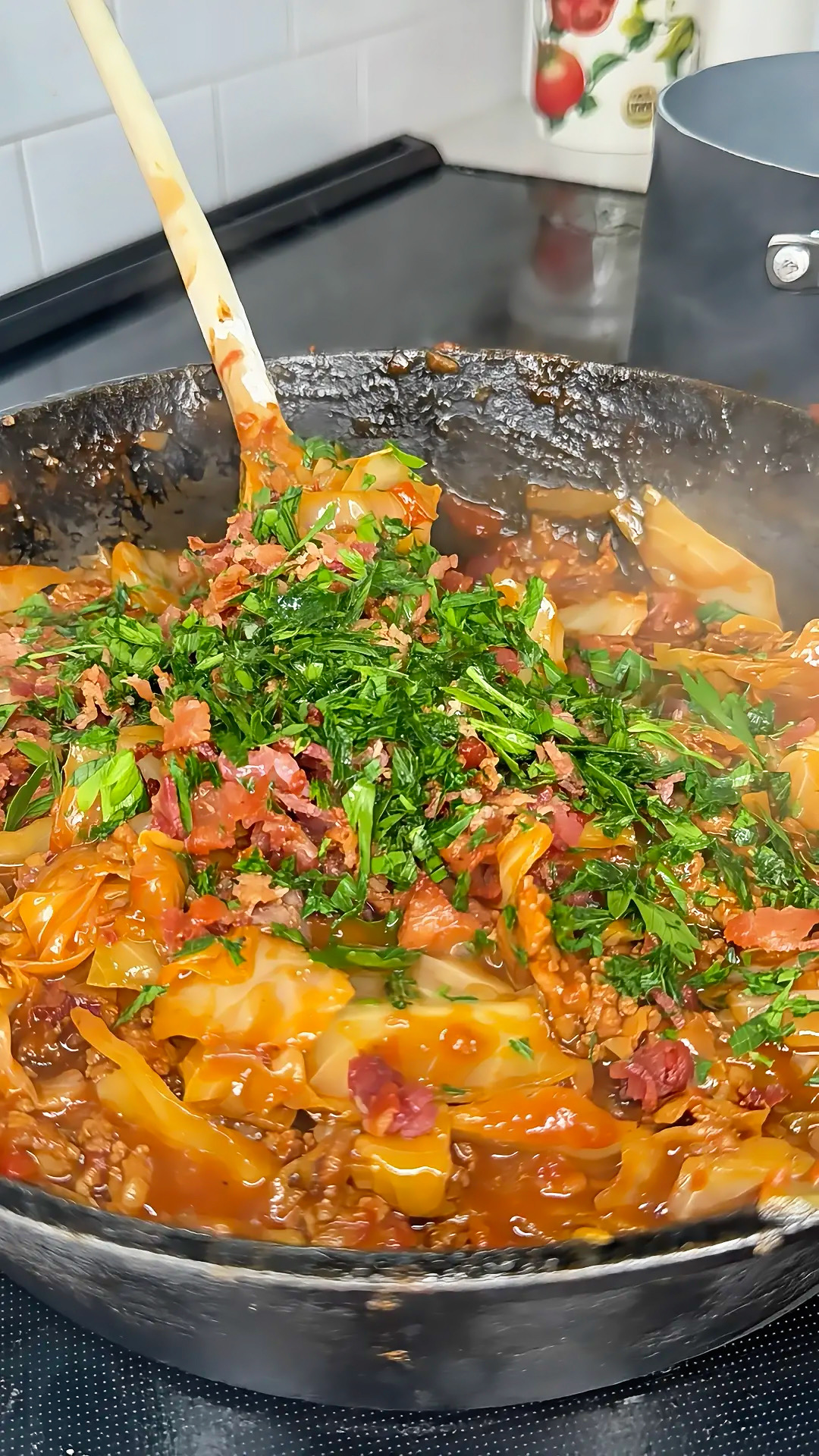 A skillet filled with a saucy mixture of cabbage, ground meat, chopped herbs, and bacon, being stirred with a wooden spoon on a stovetop. Steam rises from the hot, freshly cooked dish.