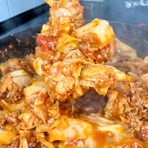 A hand holds a spatula lifting saucy stuffed cabbage rolls filled with ground meat from a steaming skillet.
