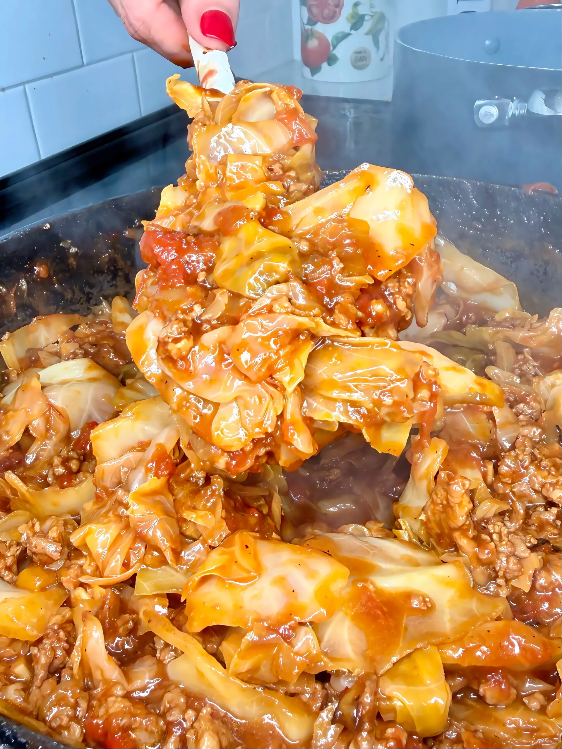 A hand holds a spatula lifting saucy stuffed cabbage rolls filled with ground meat from a steaming skillet.