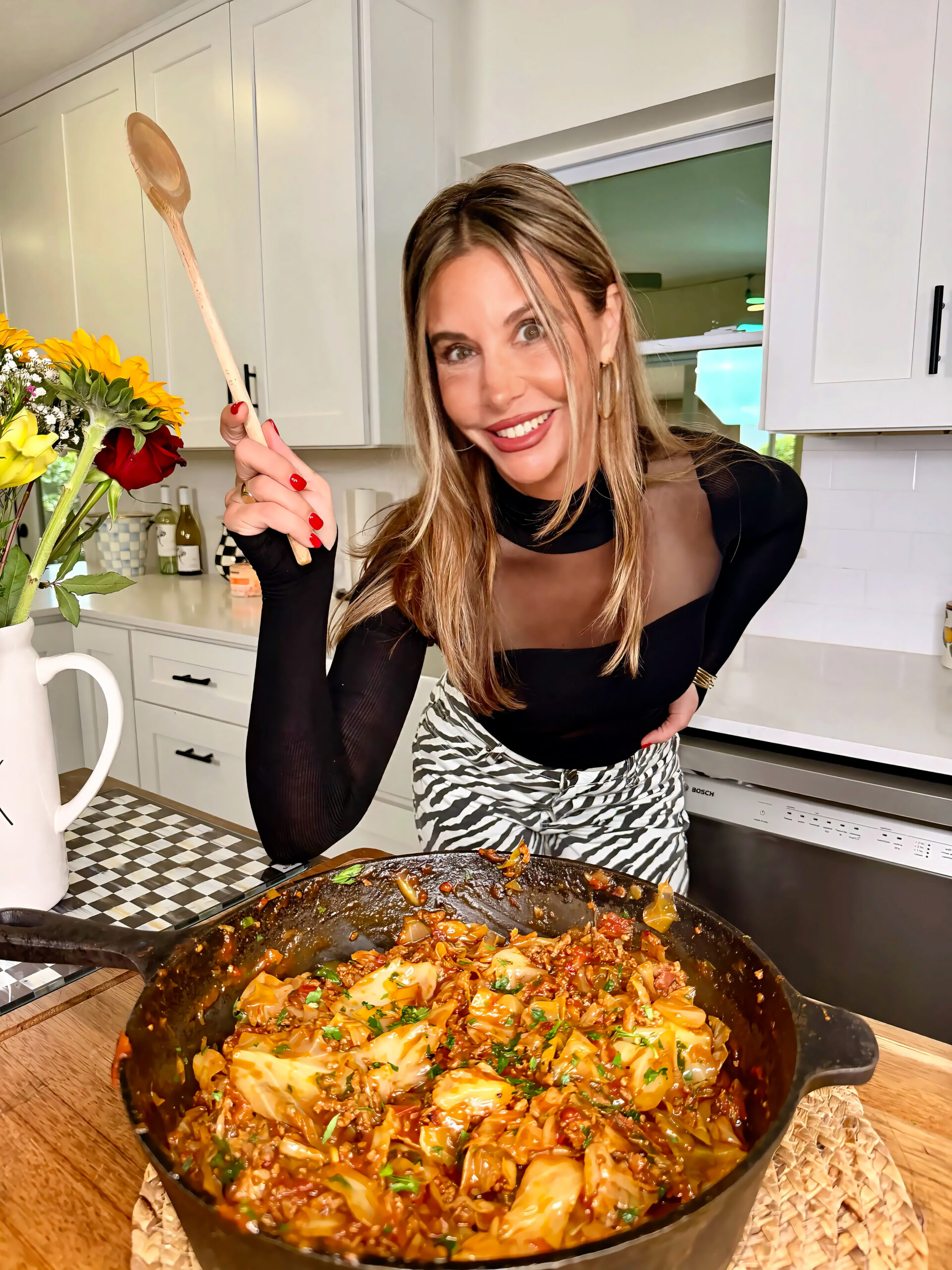 A smiling woman in a black top holds a wooden spoon next to a skillet of cooked food in a modern kitchen with white cabinets and a vase of sunflowers.