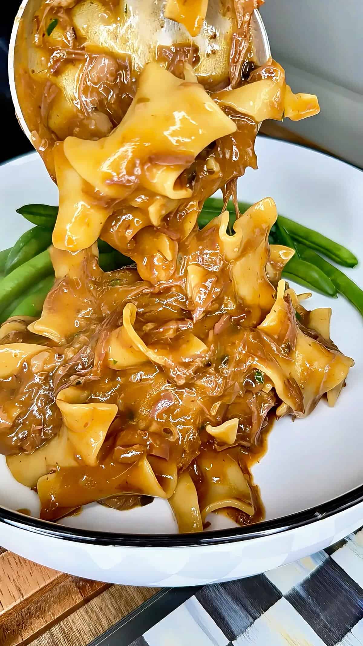 Wide egg noodles in a thick brown gravy with shredded meat are being served onto a plate with green beans.