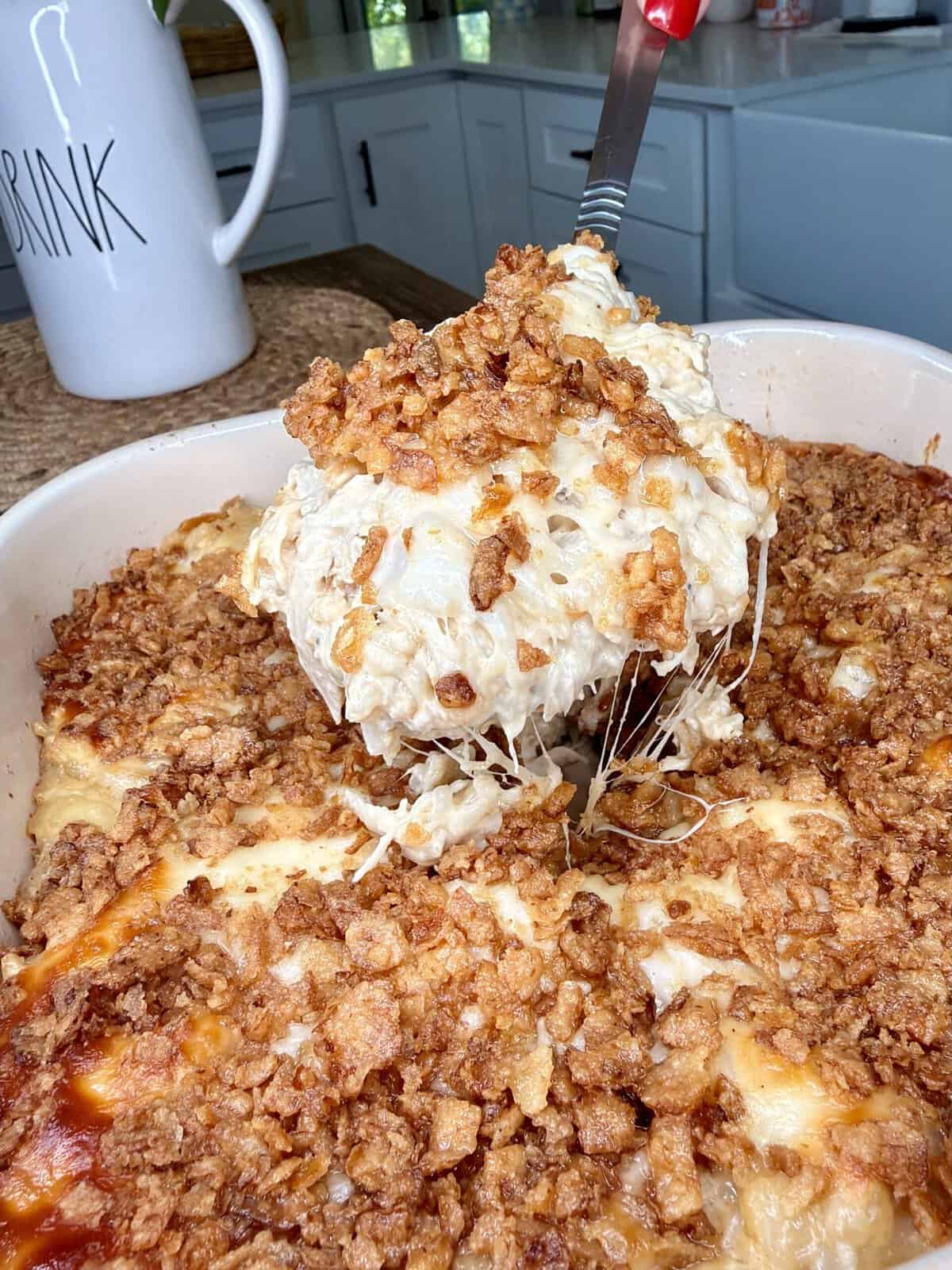 A serving of cheesy casserole with a crispy topping is being lifted from a baking dish in a kitchen. A white mug labeled DRINK is visible in the background.