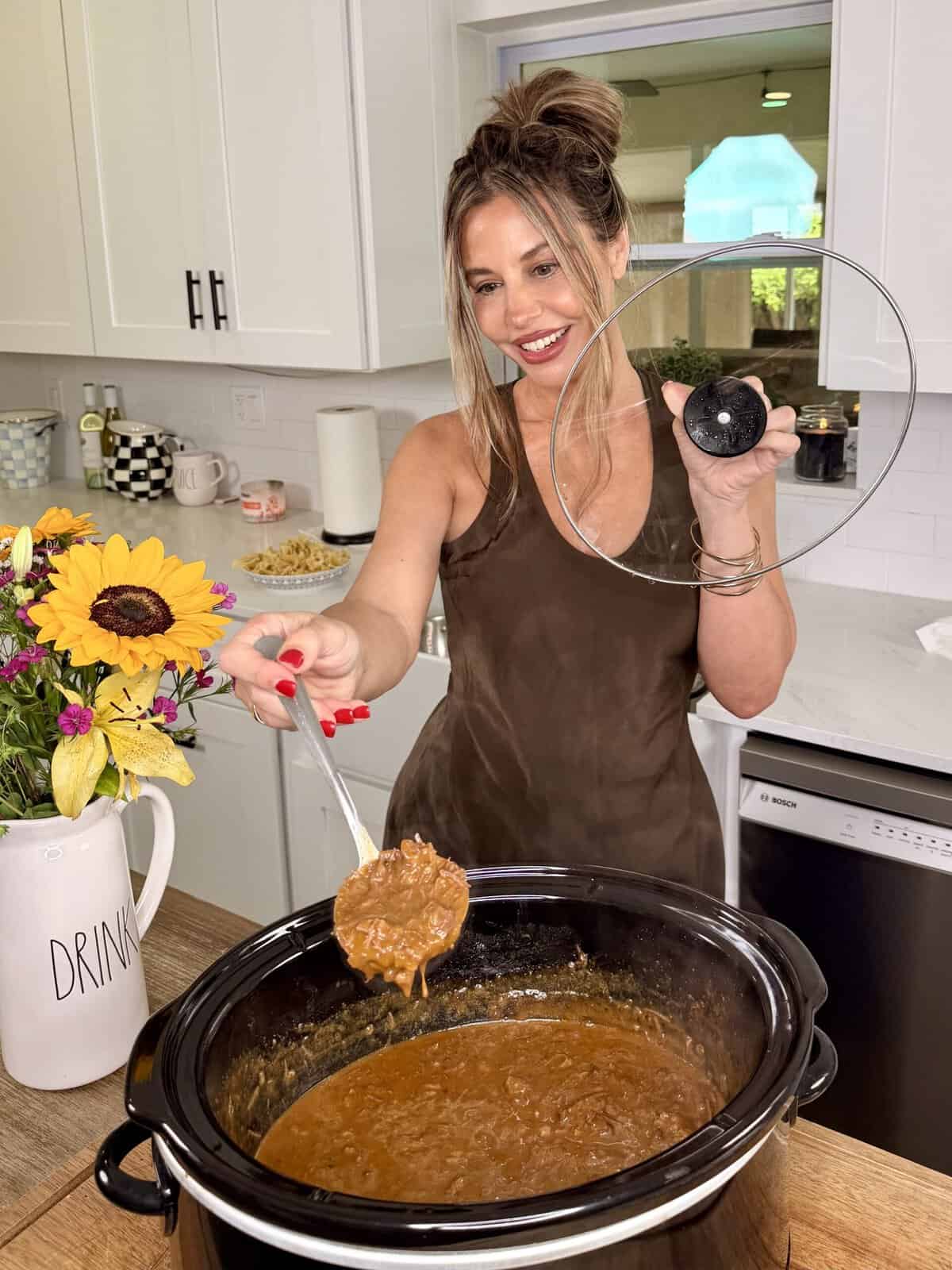 A smiling woman in a kitchen lifts the lid off a slow cooker and ladles out thick stew. A vase of sunflowers and a drink pitcher are on the counter.