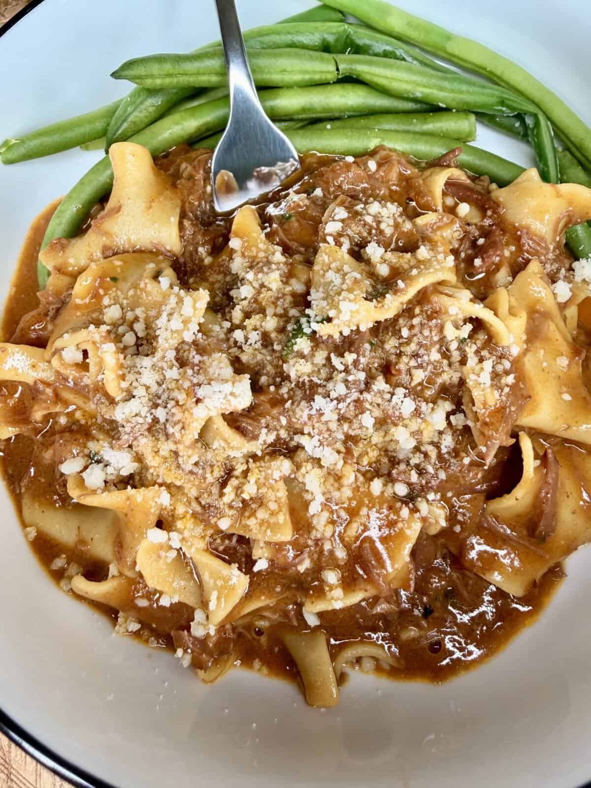 Wide egg noodles in a rich, brown meat sauce topped with grated cheese, served with a side of fresh green beans. A fork rests on the edge of the bowl.